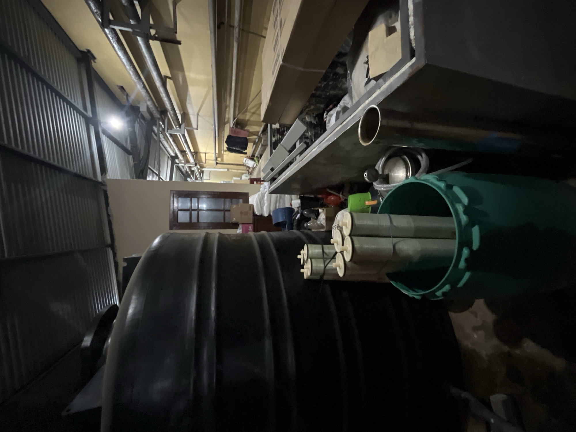 The image depicts an interior scene, possibly a storage area or workshop, with industrial elements visible. In the foreground, a large black plastic tank dominates the left side of the frame. To its right, a green plastic drum holds several cylindrical objects wrapped in plastic, tied with string. Behind these items, a long, dark rectangular structure is partially visible, filled with various objects, including cardboard boxes.
The background reveals a yellow-painted wall with a network of pipes running along it, supported by metal brackets. A window with wooden framing is visible to the left, and a door with similar framing is further back. The ceiling is constructed of corrugated metal. The overall lighting is dim, with a light source visible near the center of the frame, illuminating the pipes and ceiling. The scene appears to be located in an urban industrial or commercial setting. No specific elements within the image directly indicate a location within Pekalongan, Indonesia.