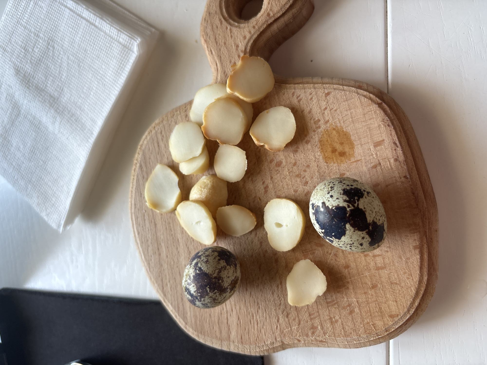 A wooden cutting board rests on a white table next to a stack of white napkins. On the cutting board are several sliced macadamia nuts and two speckled quail eggs. A black object is partially visible in the lower left corner. The location is unknown.