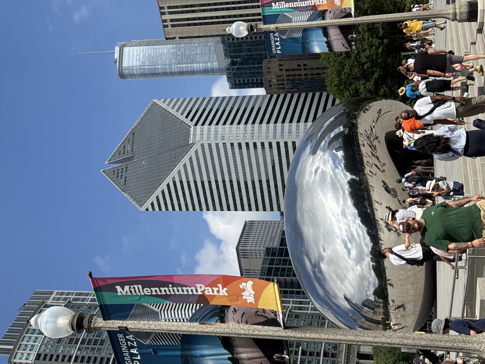 This media file captures a sunny day in Chicago's Millennium Park, featuring the iconic "Cloud Gate" sculpture, often referred to as "The Bean." The highly reflective surface of the sculpture mirrors the surrounding skyscrapers, the bright blue sky filled with scattered clouds, and the numerous people present. Several individuals are seen interacting with the sculpture, taking photos, and enjoying the park. Some are walking on the stairs and pathways around The Bean, while others are gathered near it. The scene suggests a tourist destination and a popular public gathering spot.
Notable details include the clear blue sky and bright sunlight, indicating it's likely midday. The architecture of the skyscrapers is prominent, reflecting in the sculpture, and some signage is visible. One banner reads "Millennium Park," and another smaller one beneath it says "GRAINGER PLAZA." A flag with colorful stripes, also bearing the "Millennium Park" text and a stylized figure, is attached to a lamppost. The mood appears lively and relaxed, typical of a pleasant day in a bustling urban park. The image is oriented vertically, with the sky and upper parts of the buildings appearing on the left and the ground and people on the right, as if the camera was tilted.