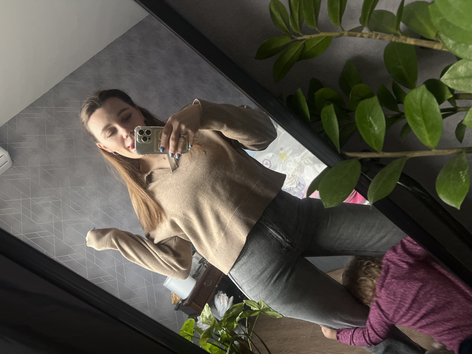 An interior scene is depicted through a mirror selfie, purportedly located in the city of Leninakan, Russia. The primary subject is an adult female, light-skinned with long brown hair, wearing a light brown long-sleeved collared sweater and grey jeans. She holds a light-colored smartphone with a clear case and a circular grip in her right hand, angled towards the mirror. Her left arm is raised with the hand near her head. She displays an expressive facial gesture with her tongue partially extended. Light blue nail polish is visible on her fingernails, and she wears rings on her left hand.
In the lower left, a young child, with light curly hair and wearing a purple long-sleeved shirt, is partially visible, clinging to the woman's left leg with both hands. The child's face is not visible.
The mirror, framed in black, reflects the interior space. The background consists of a dark grey wall featuring a subtle, reflective geometric pattern. An air conditioning unit or vent is visible near the top left of the reflection. To the left, a dark wooden dresser or cabinet is visible, holding a white cylindrical object and some green foliage. A large, leafy green plant is partially visible in the reflected lower-left corner. In the immediate foreground on the right, a Zamioculcas zamiifolia (ZZ plant) with multiple stalks and dark green leaves is prominently displayed, partially obscuring the mirror's lower frame. The lighting appears to be ambient indoor illumination.
