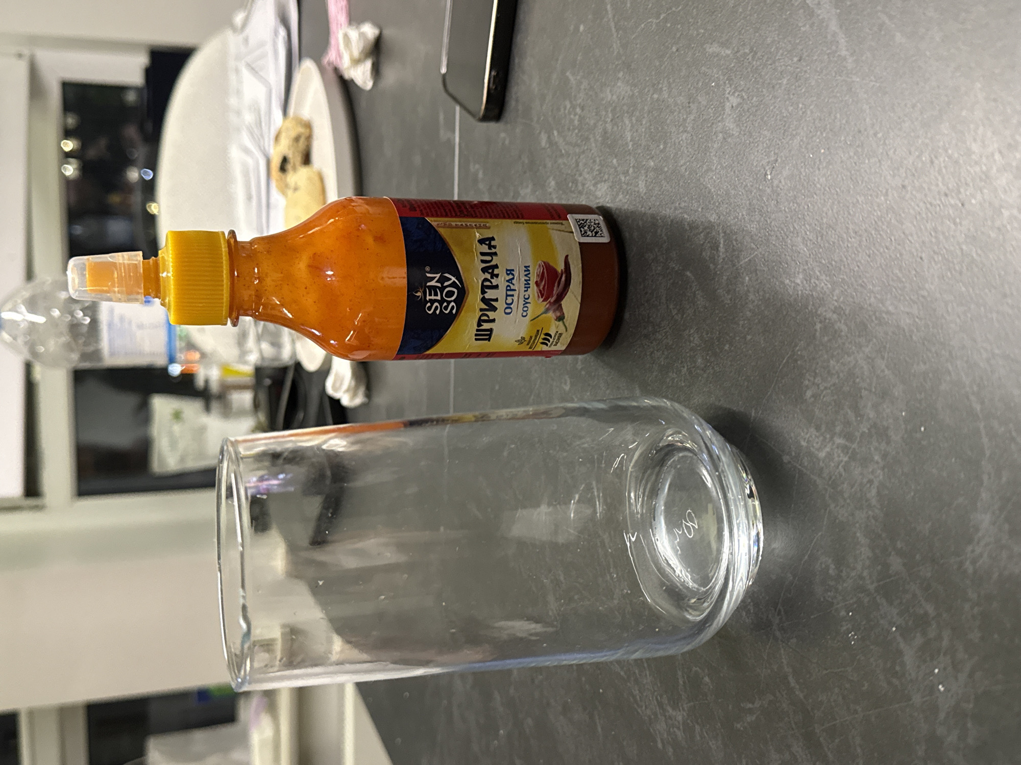 A close-up shot shows a bottle of Sen Soy Sriracha hot sauce placed horizontally next to a clear glass. The bottle is orange and red, with a yellow cap and a transparent nozzle. The label on the bottle features the Sen Soy logo and text in Russian, including "ШРИРАЧА," "ОСТРАЯ," and "соус чили." A small image of a chili pepper and a rose is also visible on the label. The glass is empty and transparent, with a thick base.
The scene appears to be indoors, likely on a kitchen counter or table, given the dark gray textured surface. In the background, out of focus, hints of what might be kitchen cabinets, lights, and a plate with food can be seen. The lighting suggests it might be evening or a dimly lit room. The overall mood is neutral, focusing on the presentation of the hot sauce and the glass. The image is captured with a shallow depth of field, blurring the background and highlighting the main subjects.