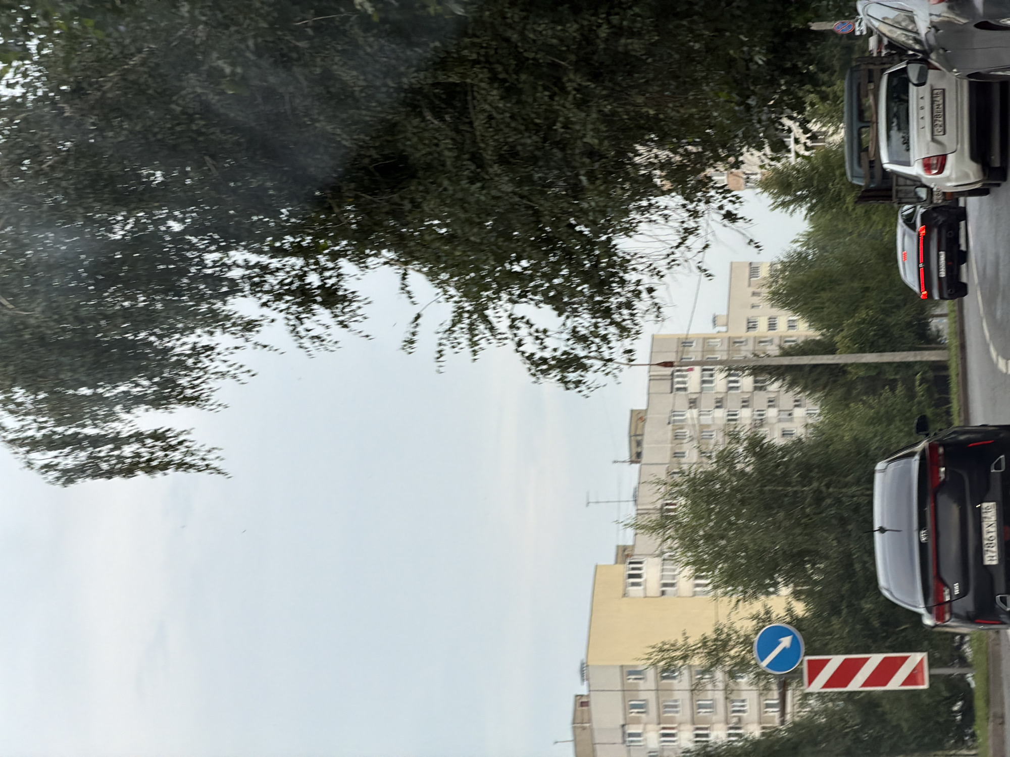 The image shows a street scene in a city, likely during the daytime under an overcast sky. In the foreground, a black Kia sedan is visible, with its rear license plate displaying the characters "H786TX 146". To the left of the Kia, there is a road sign with a blue circle containing a white arrow pointing downwards and to the left, and below it, a vertical red and white striped barrier.
In the middle ground, other vehicles are present, including a white Lada sedan with license plate "C278BM 146" and a dark truck behind it. A silver car is partially visible on the right edge of the frame.
The background is dominated by multi-story apartment buildings, some with light-colored facades and yellow accents, and others with a more uniform gray appearance. Numerous green trees are scattered throughout the scene, with a large, dense tree with dark leaves obscuring a significant portion of the upper right side of the image. The overall impression is of a residential area with traffic.