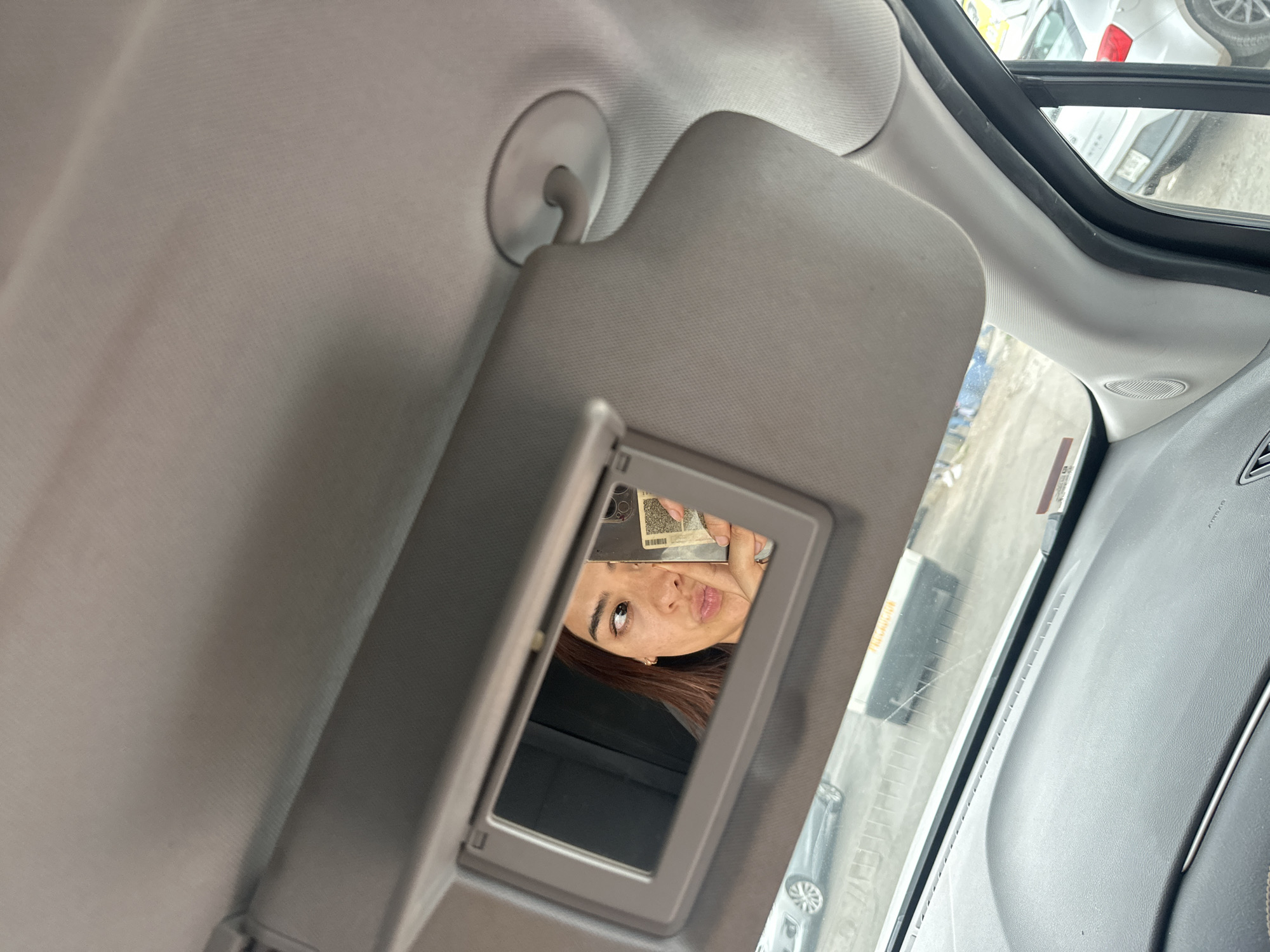 The image shows the interior of a car. The sun visor is down and open, revealing a mirror in which a woman is visible. She is holding a phone with her hand. The surrounding environment reflected in the visor shows vehicles and buildings. The context of the image indicates that the location is Puebla, Mexico.