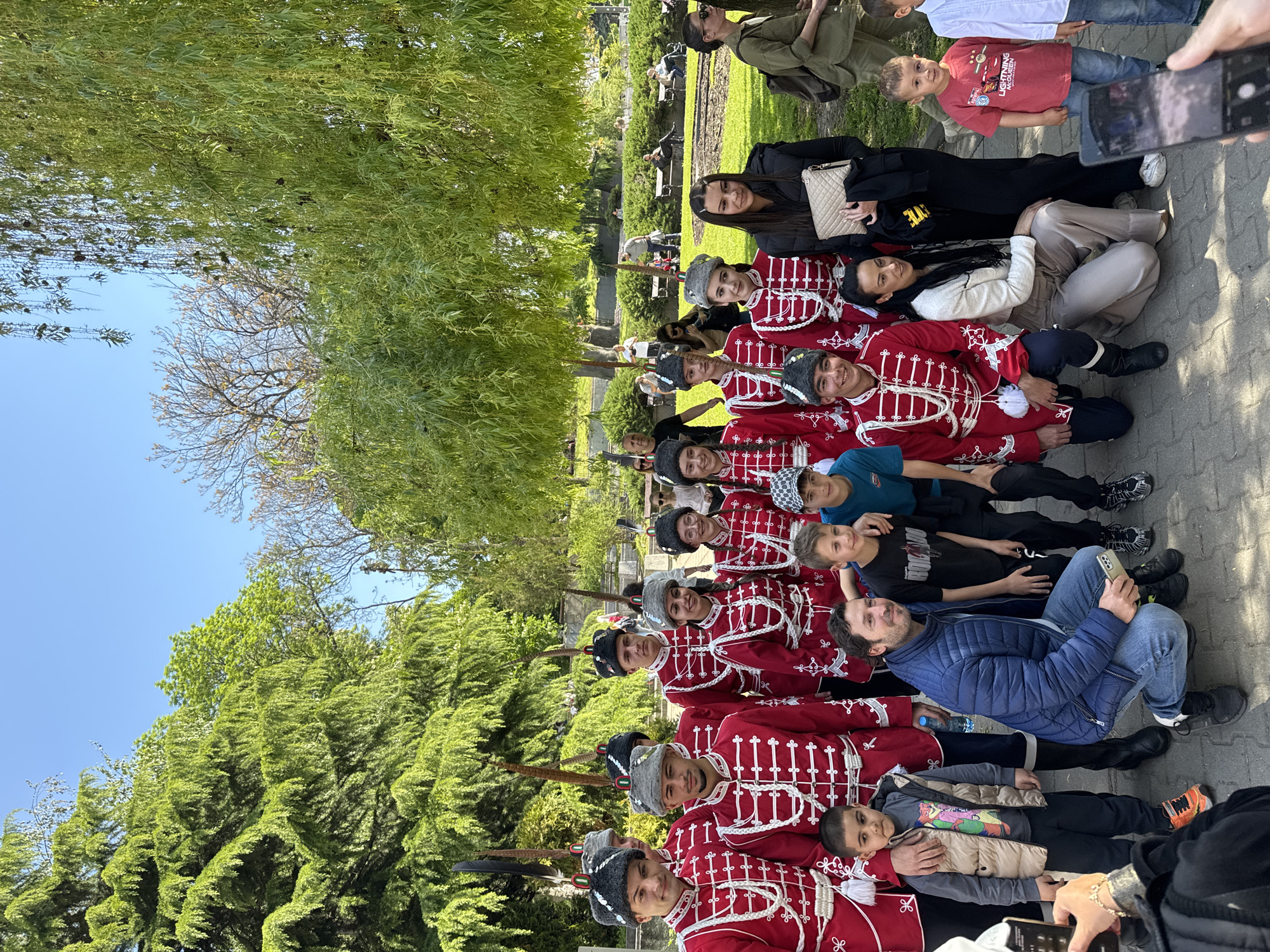 A large group of people is seated on a paved area outdoors, likely in a city park, with the sky and trees visible in the background. The location is likely in the city of Sofia, Bulgaria. The majority of the group consists of young men dressed in red and white military-style uniforms with traditional hats, presumably part of a ceremonial guard or historical re-enactment group. They are seated close together, creating a long line.
Interspersed among the uniformed men are other people, including children, women, and men dressed in casual clothing. Some of these individuals appear to be interacting with the uniformed men, possibly posing for photos or engaging in conversation. Several people in the background are also present.
The environment includes green trees, a clear blue sky, and what appears to be a park or garden setting in the background. The overall impression is that of a public event or gathering taking place in an urban outdoor setting.