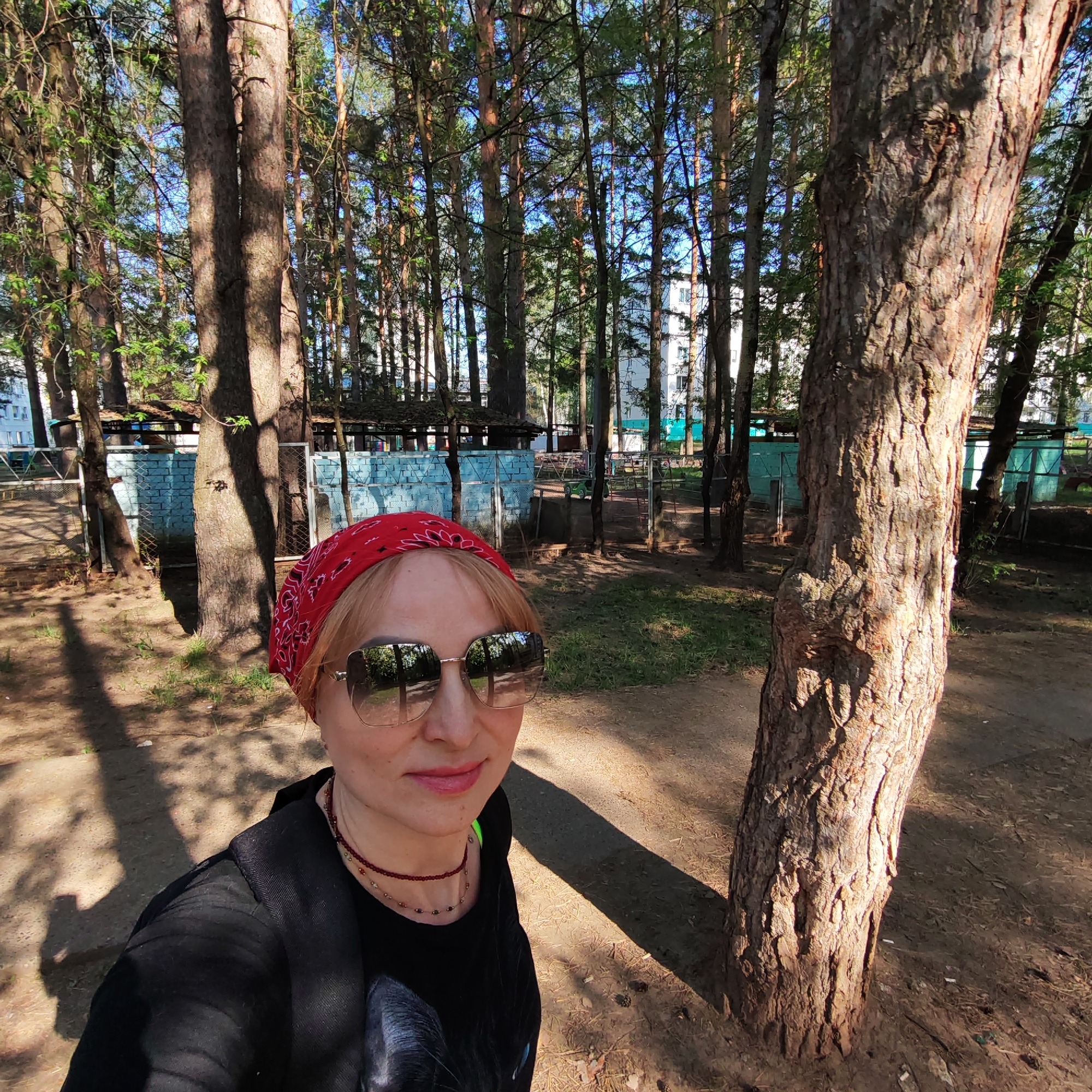 A fair-skinned woman with blonde hair and sunglasses, wearing a red bandana, stands in a park or wooded area. She is wearing a black top with a print of a black cat on it, and a beaded necklace. The background consists of tall, thin trees, some of which appear to be pines, with a building visible through the trees in the distance. There is also a low fence or structure in the background, constructed from blue painted brick or similar material. The ground is covered in dirt and fallen pine needles. The location is Chaikovsky, Russia.