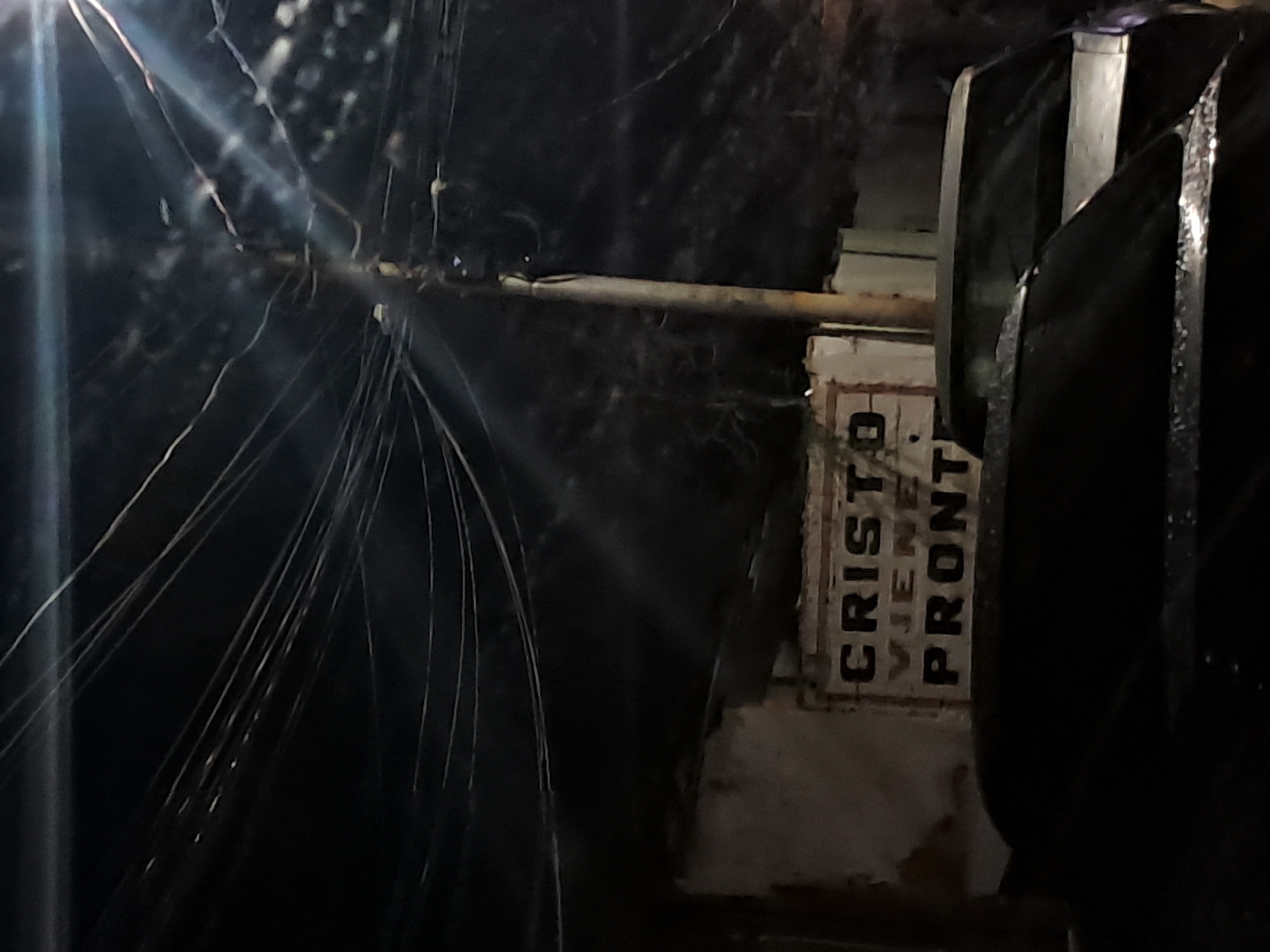 The image captures a nighttime scene in Cumana, Venezuela, with a prominent utility pole and a tangle of overhead wires dominating the upper portion. Below, a building with a weathered facade is visible, featuring a sign with the text "CRISTO VIENE PRONTO" (Christ is coming soon) in block letters. A portion of a vehicle's roof and windshield is visible in the foreground, suggesting its presence on a street or in a parking area. The overall lighting is dim, with artificial light sources creating lens flares and illuminating parts of the scene. The environment appears to be an urban setting, characterized by the presence of electrical infrastructure and buildings.