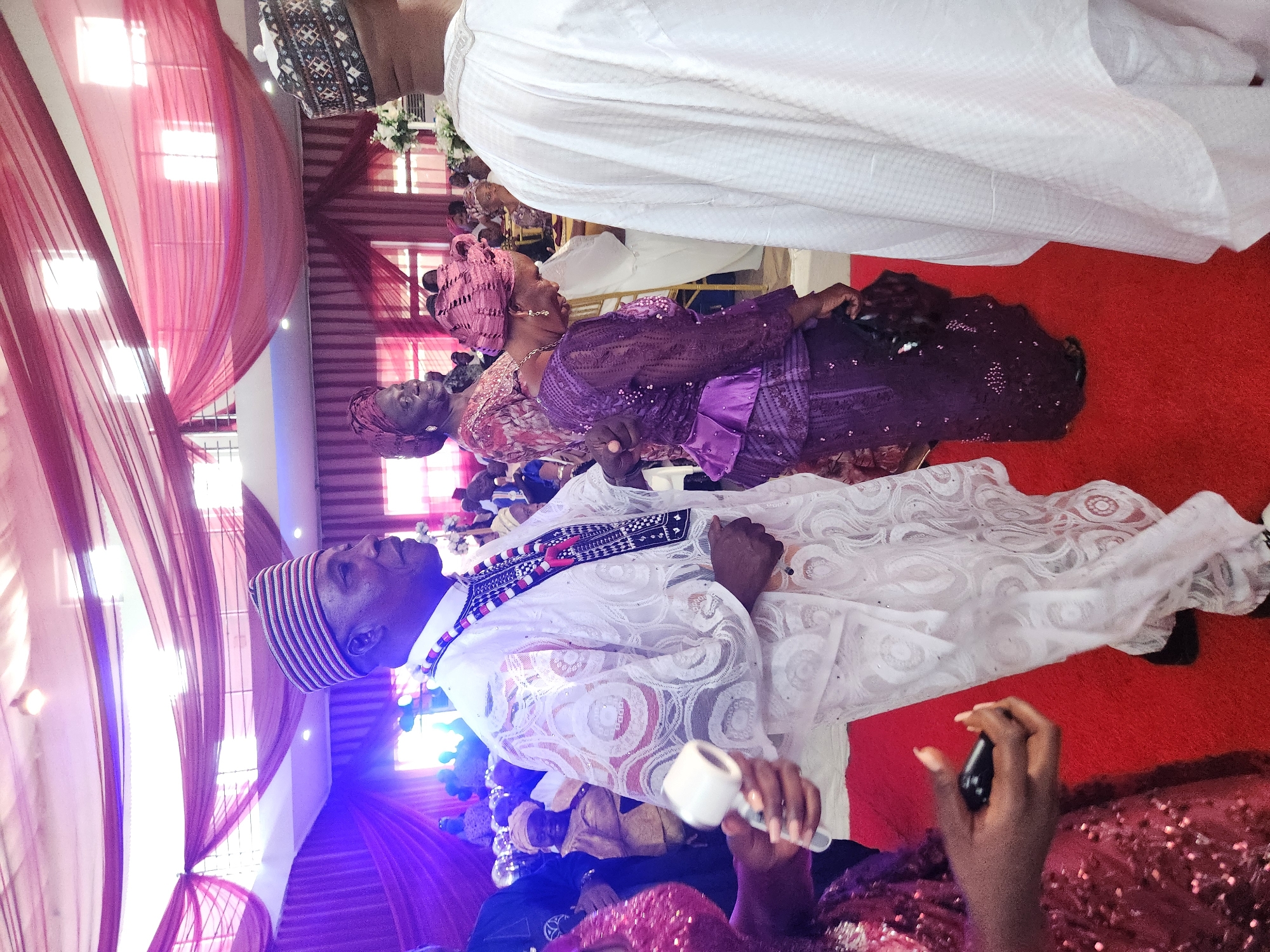 The image captures a gathering of people, likely at a social event such as a wedding, in Barden Yana, Nigeria. The scene is set indoors, with a red carpet covering the floor and draped fabric in shades of red and pink adorning the ceiling and walls, suggesting a festive atmosphere.
In the foreground, a man in traditional Nigerian attire, a white agbada with intricate lace detailing and a striped cap, is the central figure. He wears a colorful beaded necklace. To his left, a woman in a sequined red dress is partially visible, holding what appears to be a small white object in her right hand and a black object in her left. To the man's right, another man, also in white traditional attire and a cap, is seen from the back.
In the mid-ground, a woman in a purple lace outfit and a matching headscarf is looking down, with a beaded necklace visible. Behind her, other guests are milling about, some in traditional clothing. The background reveals more of the venue's decor and additional attendees, with glimpses of white chairs and floral arrangements. The lighting appears to be a mix of ambient and possibly colored stage lighting, casting a purple hue in some areas. The overall impression is one of a lively celebration.