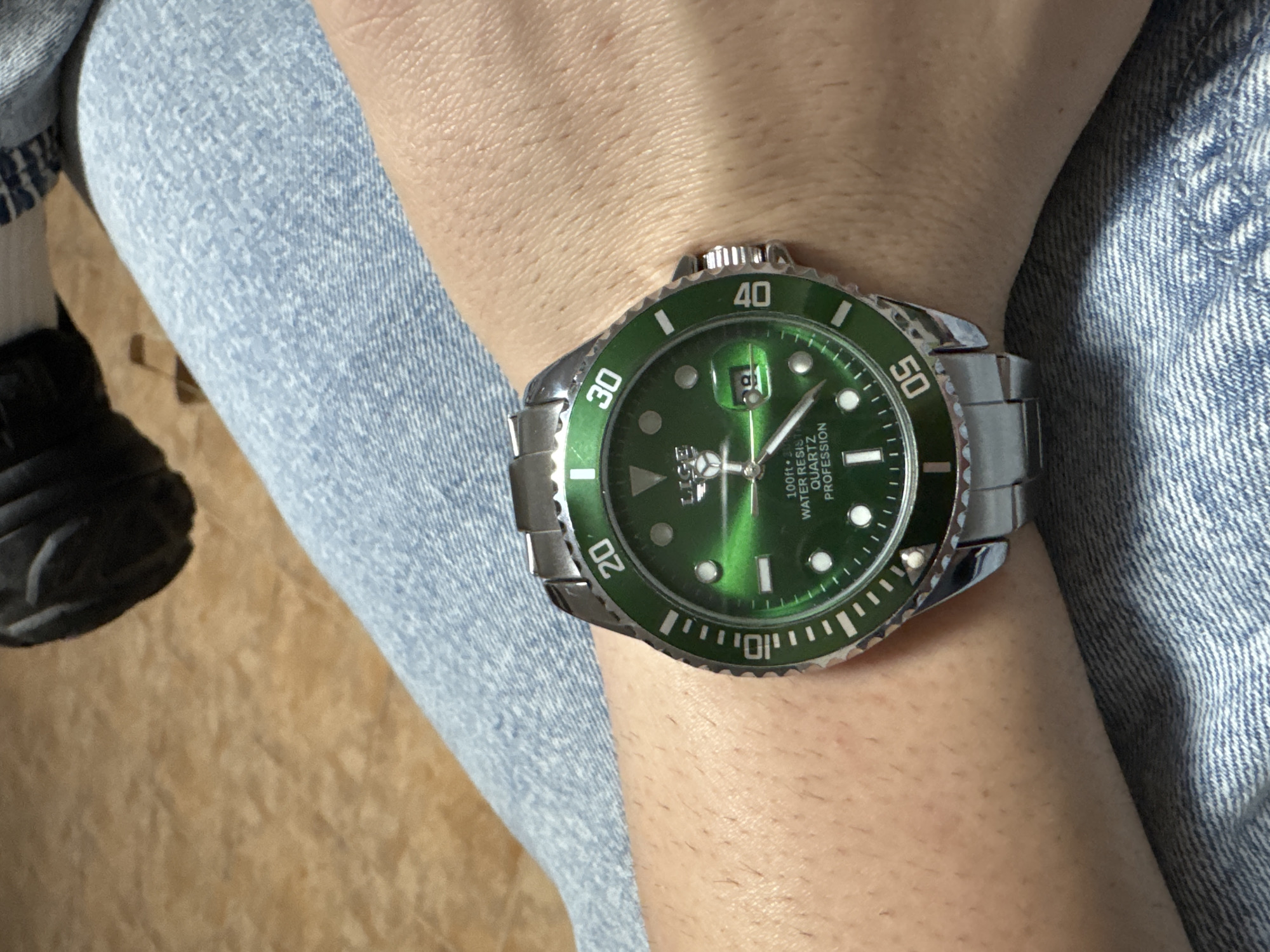 A close-up, high-angle shot depicts a person's light-skinned forearm wearing a wristwatch, resting on a leg clad in light blue denim jeans. The watch is a LIGE brand model with a silver-toned metal case and bracelet, featuring a prominent green sunburst dial and a green unidirectional rotating bezel with silver markings. The dial displays the brand name "LIGE," three hands (hour, minute, second), a date window at the 3 o'clock position showing the number "19," and text indicating "100ft WATER RESIST QUARTZ PROFESSION." Hour markers are applied dots and rectangles. The skin of the forearm shows visible fine hair. In the background, a blurred light brown surface, possibly a wooden or laminate floor, is visible. A dark, out-of-focus object, potentially part of footwear, is present in the upper left corner of the frame.