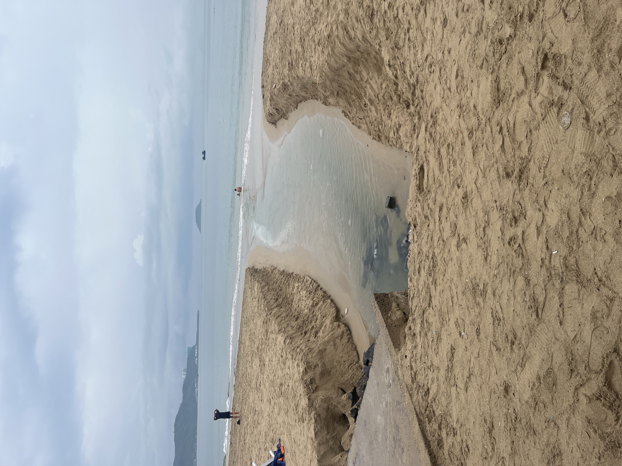 The image captures a serene coastal scene at Bac Nha Trang Ward, Vietnam, under an overcast sky. The primary subjects are the expansive sandy beach and the calm, light blue-green sea. A small, natural channel of water flows across the sand, creating a meandering path into the ocean.
In the water, near where the channel meets the sea, two individuals are visible; one appears to be an adult, and the other, smaller, is likely a child, both possibly bathing or wading. Another person stands on the sandy shore to the left, dressed in dark clothing, looking out towards the sea. Several boats or ships are present in the distant waters, suggesting maritime activity.
The beach itself is characterized by light brown sand, showing numerous footprints and tracks. A discarded plastic cup or lid is visible in the sand on the right side. In the water of the flowing channel, a dark, rectangular object is partially submerged. Mountains or hills rise in the background on both sides of the distant horizon. The weather is cloudy and calm, contributing to a peaceful atmosphere. No visible text is present in the image.