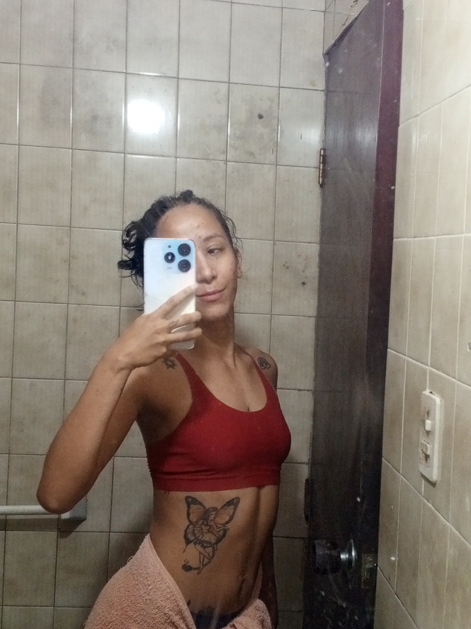 A person with dark hair pulled back, wearing a red sports bra, stands in a bathroom and takes a selfie using a white smartphone. The person has tattoos on their shoulder, hand, and torso. A towel is wrapped around their waist. The bathroom has light-colored square tiles on the walls. A dark, wooden door with a silver doorknob is on the right side of the frame. A light switch is visible on the tiled wall near the door. The environment appears to be a bathroom in a private residence. It is not possible to identify the location in Porlamar, Venezuela without additional information.