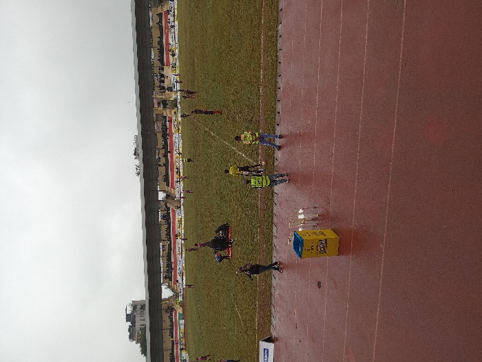 The image displays Kinoru Stadium in Meru, Kenya, under an overcast sky. The foreground features a wet, reddish-brown synthetic running track with visible white lane markings. Positioned on the track are a yellow rectangular waste bin with "Safaricom" branding and a blue top, alongside two white plastic chairs. Three individuals are present near the track: one person in dark clothing walks left, and two individuals wearing yellow-lime green reflective vests stand at the edge of the adjacent football pitch.
The football pitch itself is heavily waterlogged, muddy, and exhibits numerous patches of bare earth, with obscured white line markings. Multiple individuals, identified as football players wearing dark-colored jerseys, are distributed across the field. A cluster of 3-5 players and other personnel are gathered on the left-mid section of the pitch, with some individuals bending or kneeling. A single player in a yellow jersey is visible on the right side of the field.
In the background, a multi-tiered grandstand with a dark, possibly corrugated metal roof spans the width of the image. The grandstand holds a moderate number of spectators, and advertising banners are visible along its lower railings. Above and to the left of the grandstand, a multi-story urban building with a flat roof is discernible.
