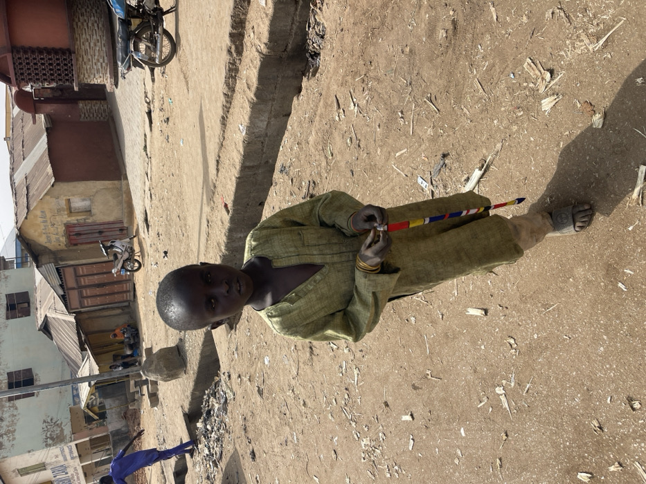 The image depicts a young, dark-skinned child standing centrally on an unpaved, dusty ground. The child wears a loose-fitting, long-sleeved olive-green garment, light-colored trousers, and dark sandals. Multiple gold-colored bangles are visible on their left wrist. In their hands, the child holds a vertically oriented object wrapped with multicolored fabric (red, yellow, blue, white). The child looks directly at the camera with a neutral expression.
To the child's left, a person in a blue outfit is partially visible, standing near a pile of debris in a ditch or gutter. Beyond them, a building with a light-colored facade and a partially visible sign that reads "BUILDING ZARIA" is present. Several other structures with corrugated metal roofs and varying wall finishes are visible in the background, along with a white motorcycle parked to the left.
To the child's right, a concrete curb or low wall runs horizontally across the mid-ground, separating the foreground's dirt path from a slightly elevated, possibly paved, area. More buildings with corrugated roofs and a parked motorcycle are visible further back. The ground throughout the scene is unpaved, covered in dirt, and scattered with debris including wood fragments and trash. Strong sunlight casts clear shadows. The specific location is identified as Zaria, Nigeria, based on the visible signboard.