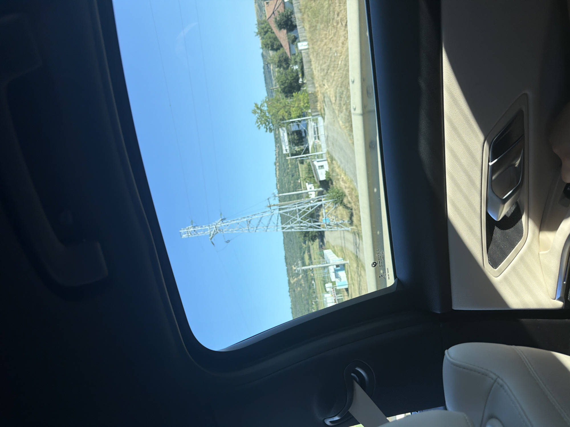 The image is taken from inside a vehicle, looking out the side window. The view outside is a rural landscape under a clear blue sky. Dominating the scene is a tall, metal electricity pylon. In the middle ground, there are some buildings, including a small white shed and a larger structure with a blue accent. A dirt road leads towards these buildings, passing through dry, grassy fields. Further in the background, rolling hills are visible, covered in vegetation. A few houses with red-tiled roofs are scattered throughout the landscape. A white fence lines the area around one of the properties. The car's interior is partially visible, showing a cream-colored seat and a section of the door panel with a silver handle and speaker grill. The photo appears to have been taken on a sunny day. The presence of the electricity pylon and the general rural setting suggest a location outside of a major urban center. Considering the prompt's mention of Zorleni, Romania, this image depicts a view consistent with the outskirts or surrounding countryside of such a town.