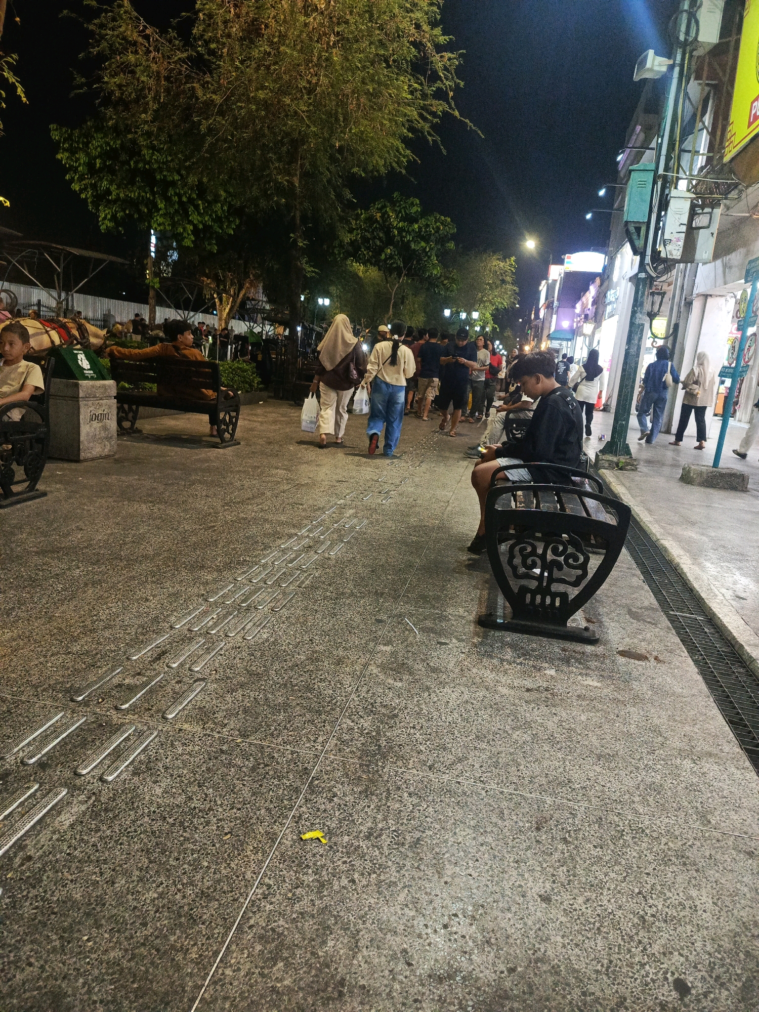 The image depicts Malioboro Street in Yogyakarta, Indonesia, at night. A young man in a black shirt and patterned shorts is sitting on a decorative black bench on the sidewalk, looking at his phone. To his left, another person is sitting on a similar bench, looking away from the camera. In the background, a group of people are walking along the street, illuminated by streetlights and shop signs. Some individuals are wearing headscarves. The sidewalk is paved with textured tiles, and a metal grate runs along the edge of the street. Trees with dense foliage line the left side of the street. The overall atmosphere is that of a busy night market or shopping street.
