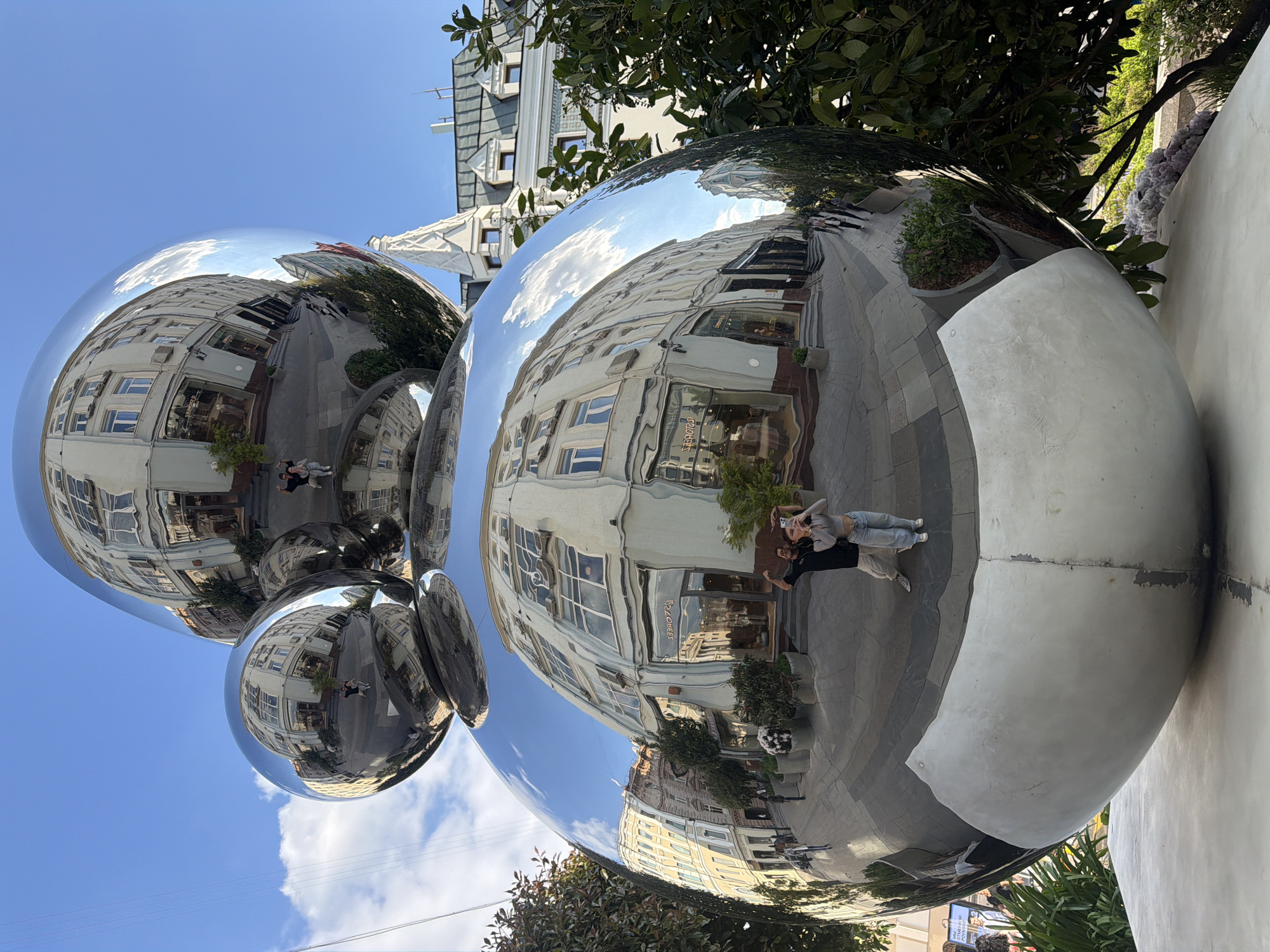 The image captures a mirrored sculpture in the city of Moscow, Russia, likely a tourist attraction or public art installation. The sculpture consists of several large, shiny, metallic spheres clustered together. The spherical surfaces reflect the surrounding urban environment, including buildings with windows, a sidewalk, trees, and the blue sky with clouds. Two people are visible in the reflection, seemingly taking a photo of themselves with the sculpture. The base of the sculpture appears to be a light-colored material. The photograph is taken from a slightly low angle, emphasizing the size and reflective properties of the spheres.