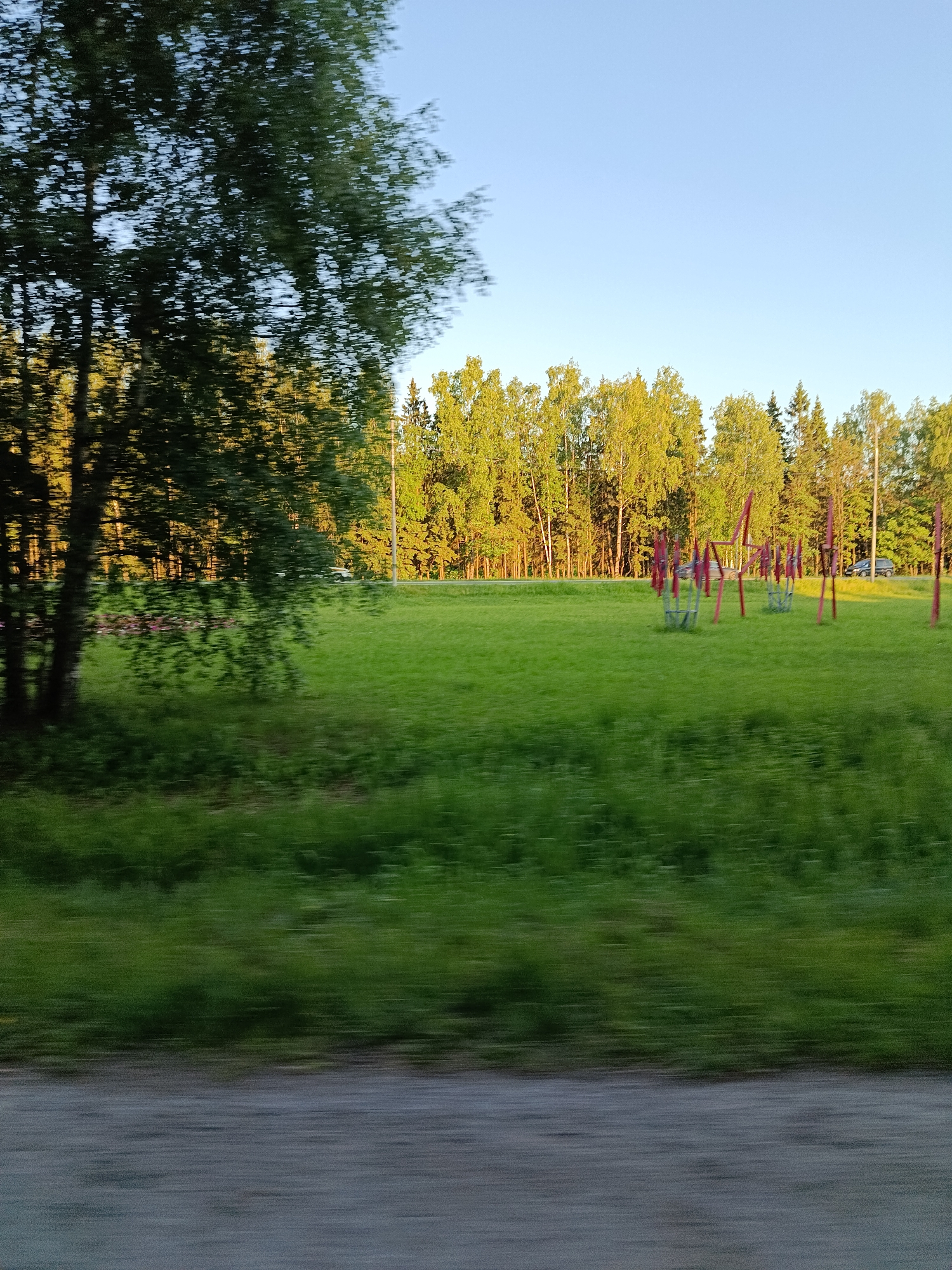 The image shows a motion-blurred view of a park-like area with a playground. The sun is setting, casting a warm glow on the trees in the background, which appear to be a mix of deciduous and coniferous. The sky is clear and blue. In the foreground, there is a grassy field with some playground equipment, primarily red metal structures that may be swings or climbing frames. The motion blur suggests the image was taken from a moving vehicle. There are no people clearly visible in the image due to the blur. The scene depicts an outdoor recreational space likely located in a suburban or park area.