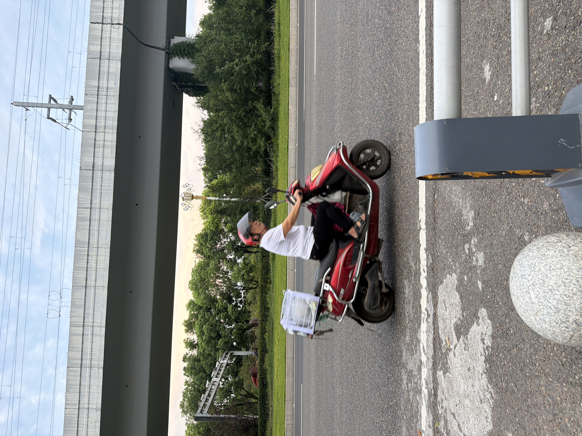 A woman wearing a helmet and a white t-shirt is riding a red scooter on a paved road in Jiangjin, China. She is carrying a white cage on the back of her scooter. In the background, there is a large elevated concrete structure, trees, and power lines. The sky is overcast with a hint of sunset light. The road has white lane markings and some patches of rough asphalt. In the foreground, there is a granite bollard and a grey barrier with yellow reflective markings.