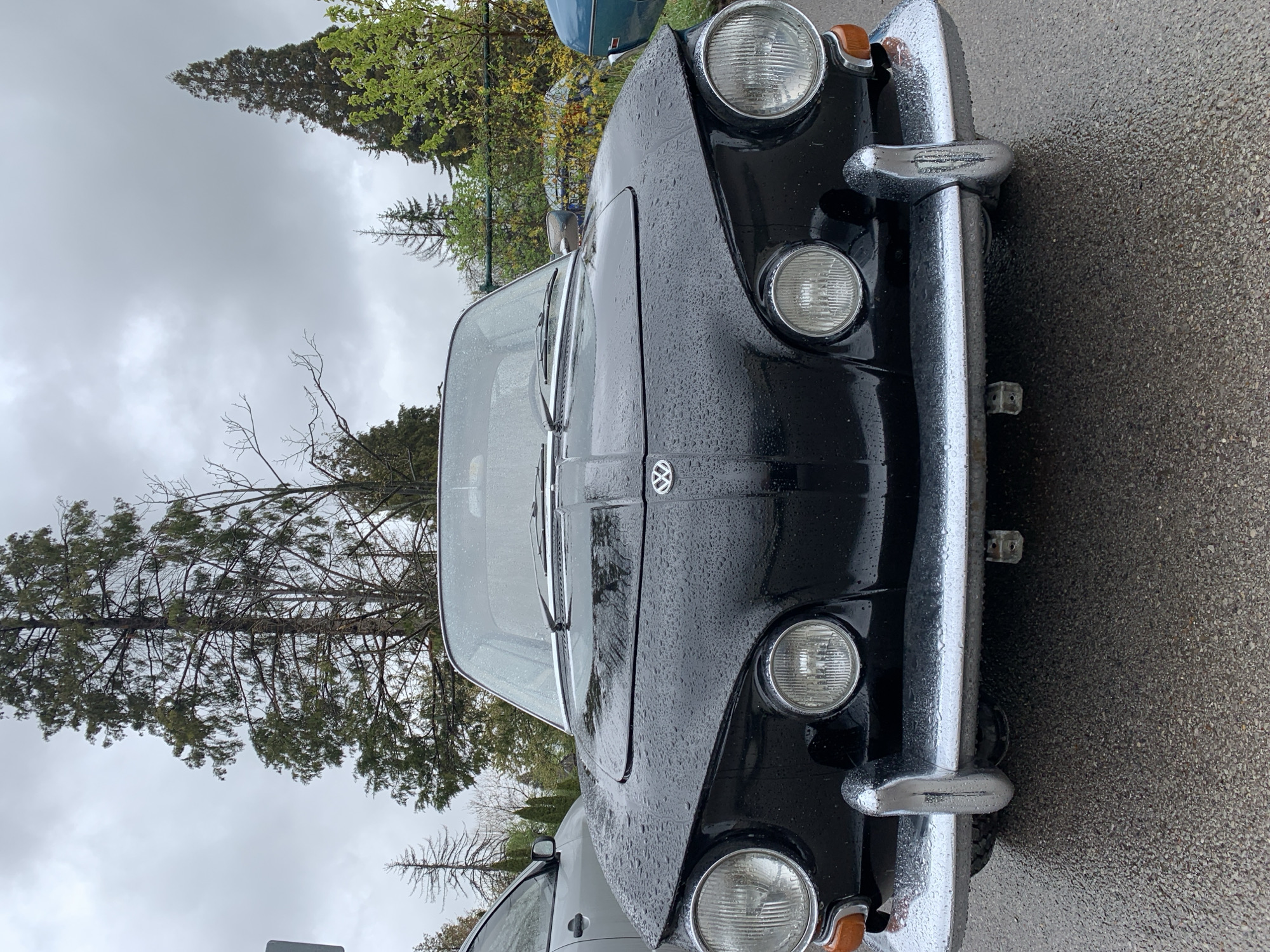 This media file captures the front view of a black, vintage Volkswagen Type 3 car, likely a Fastback or Squareback model. The car is heavily covered in water droplets, indicating it has recently rained or is currently raining. Its shiny black paint contrasts with the reflective chrome bumper, which also glistens with water. The car features prominent circular headlights, amber turn signals positioned below them, and a distinctive Volkswagen (VW) emblem on the center of the hood.
The car is parked outdoors on an asphalt or paved surface. In the background, a natural setting is visible under an overcast, cloudy sky. Various trees with both lush green foliage and some bare branches line the background, suggesting a time of year other than full summer or deep winter. A lighter-colored vehicle, possibly grey or silver, is partially visible behind the black Volkswagen on the right side of the frame. A small blue object, possibly another vehicle or a sign, can be seen in the distant left background.
No specific activity is taking place; the car appears to be stationary. The time of day is likely daytime, but the overcast conditions provide a soft, diffused light. The location context provided is Sofia, Bulgaria. Other than the Volkswagen logo, no visible text is discernible in the image.