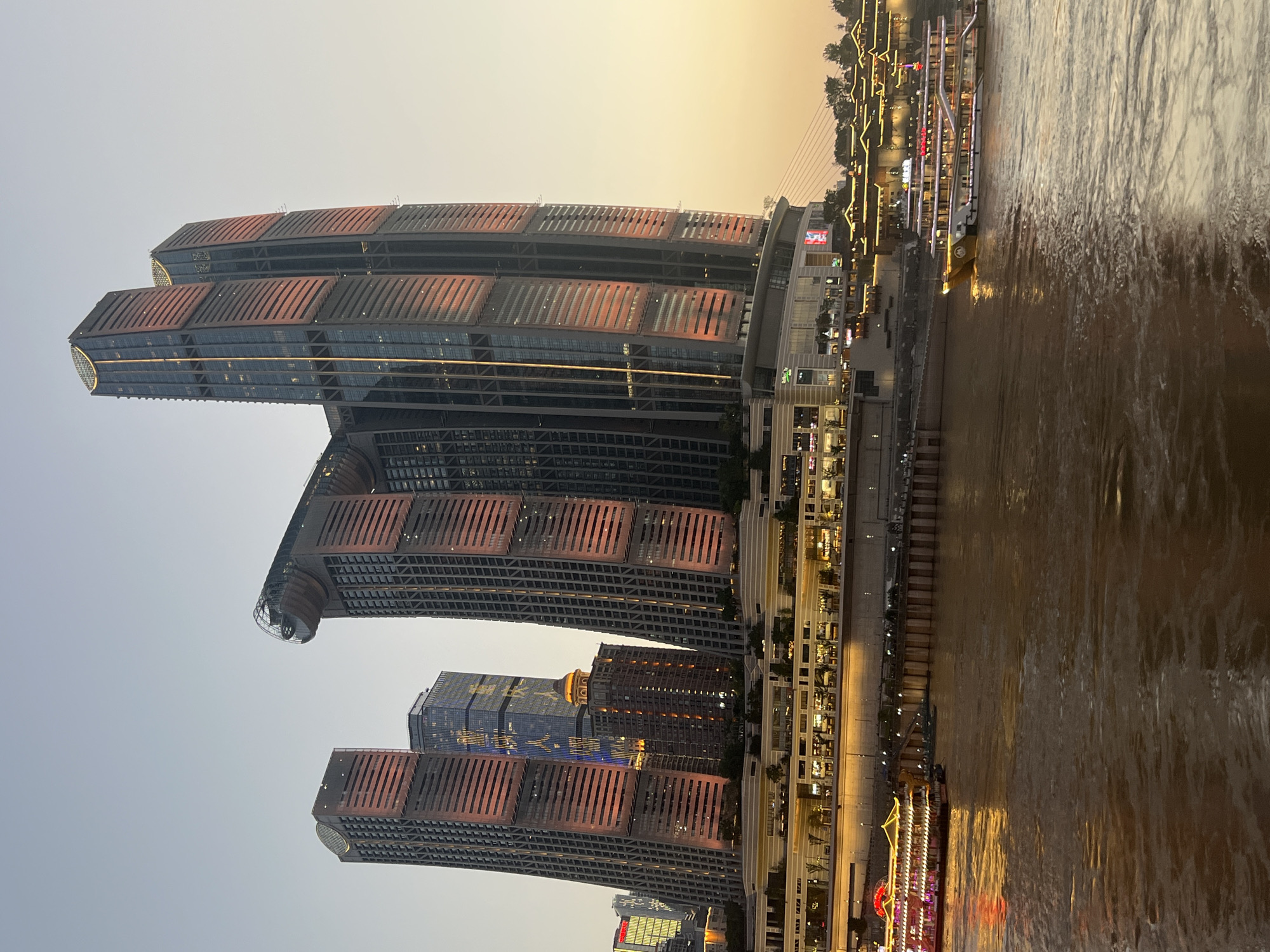 The image shows a waterfront scene in Nan'an District, China, at dusk. The sky is a pale orange and yellow, indicating either sunrise or sunset. Dominating the background are several modern skyscrapers with illuminated windows and facades. The architecture of these buildings is characterized by sharp lines and geometric shapes, with some featuring distinctive curved or angular elements. One prominent building appears to have a circular structure near its top, connected by a bridge-like element.
In the middle ground, a multi-level complex of buildings lines the waterfront. These buildings have a more traditional architectural style, with ornate roofs and numerous small, illuminated windows. The complex is brightly lit, with strings of yellow lights accentuating its features. A promenade or walkway is visible in front of this complex, with a visible railing.
The foreground is occupied by a wide river with brown, murky water. The water's surface reflects the lights from the buildings and boats, creating streaks of gold and orange. Two large passenger boats are visible on the river. The boat on the left is decorated with red and yellow lights, and appears to be docked or moving slowly. The boat on the right is also illuminated and is positioned further into the river, suggesting it is actively cruising. The riverbank is lined with a concrete embankment and what appears to be a docking area with bollards and a pier. Trees are visible in the distance along the top of the embankment. No people are clearly discernible in the image.