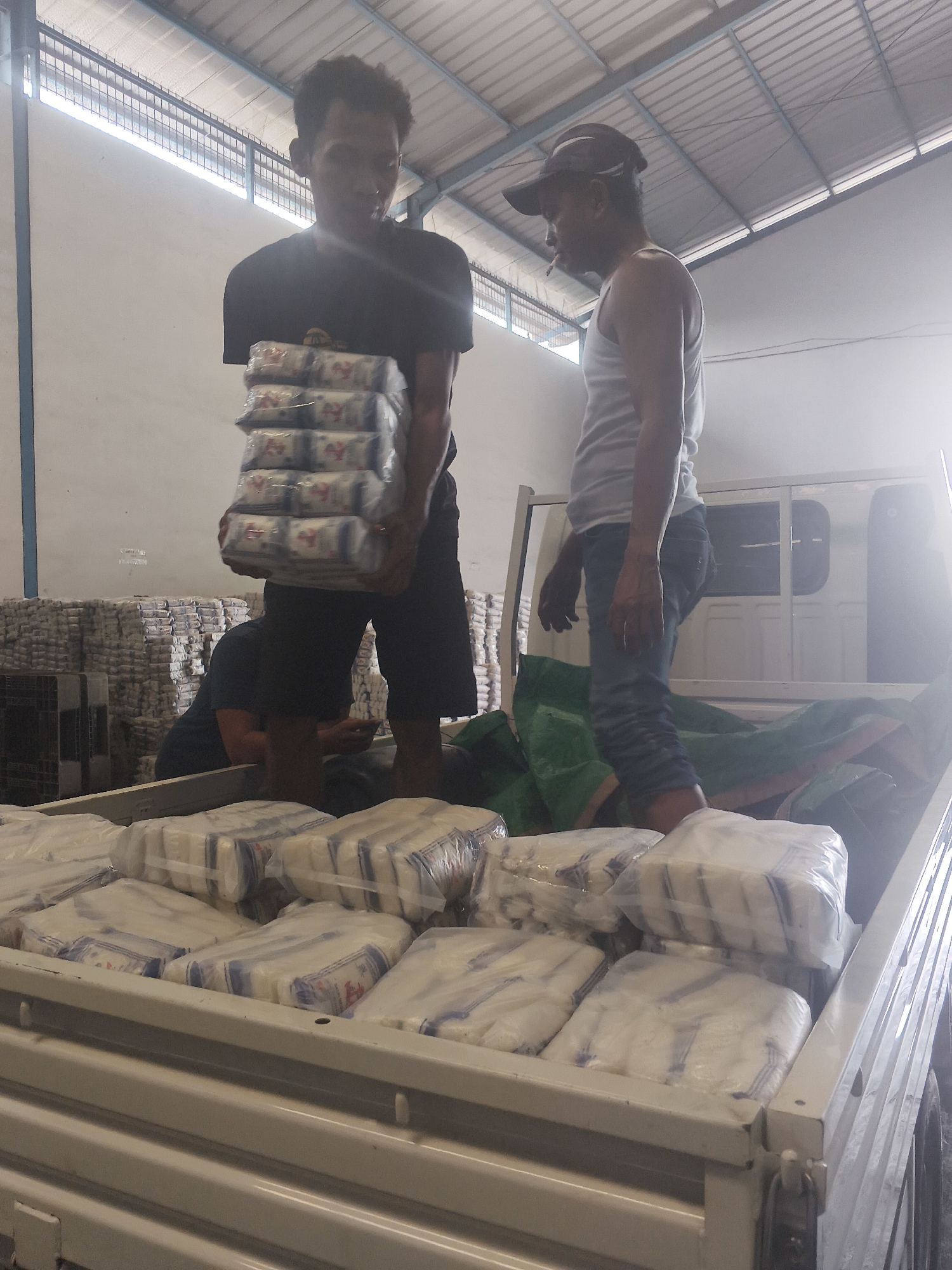 Two men are loading packaged goods into the bed of a white pickup truck at a warehouse in Bandar Lampung, Indonesia. The man on the left, wearing a black t-shirt and dark shorts, is carrying a stack of approximately six packages. The man on the right, wearing a white tank top, blue jeans, and a cap, is standing in the truck bed, holding a green tarp. The truck bed is filled with many more packages wrapped in clear plastic with blue and white labels. In the background, stacks of similar packaged goods are visible, as are the corrugated metal roof and lighting fixtures of the warehouse. The lighting suggests it is daytime.