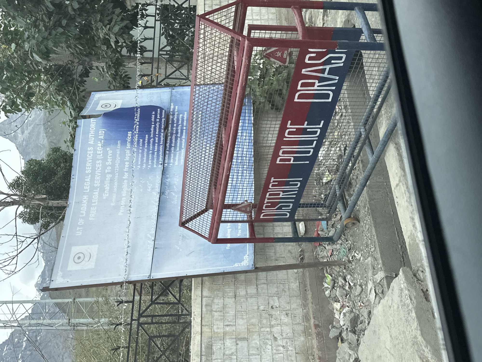 The image captures a scene in Dras, India, featuring a street-level view of a roadside barrier and a large informational sign. The barrier, painted red and blue, displays the words "DISTRICT POLICE DRAS" in white capital letters. A small red stop sign with a skull icon is affixed to the barrier. Behind the barrier, a large blue and white sign is partially visible, containing text in English and what appears to be Urdu. The English text includes "U.T OF LADAKH LEGAL SERVICES AUTHORITY FREE LEGAL SERVICES (LEGAL AID)" and lists categories of individuals eligible for free legal services. The environment appears somewhat disheveled, with debris scattered around the base of the sign and barrier. In the background, a mountainous landscape is visible, suggesting the mountainous terrain of the region.