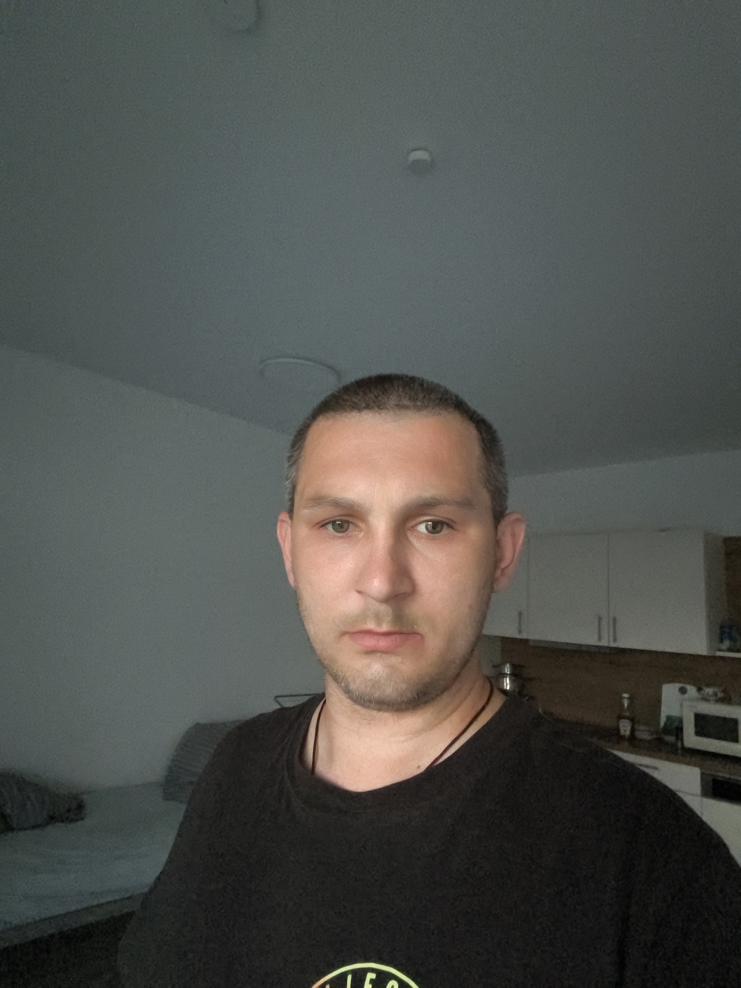 A man with short brown hair and light green eyes is looking directly at the camera in a medium shot. He is wearing a black t-shirt with a faint circular design visible on his chest. The background shows a bedroom with a grey bed and pillows on the left, and a kitchen area with white cabinets, a microwave, and a bottle of ketchup on the right. The lighting is dim, and the overall setting appears to be an apartment. The image was taken in Wolgast, Germany.