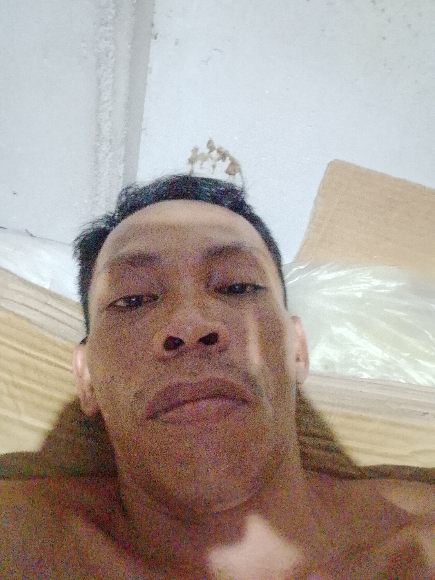 A medium close-up, low-angle selfie of a middle-aged Asian man with dark, short hair and brown skin. The man is facing the camera with a neutral expression. His shoulders and chest are visible, suggesting he might be shirtless. The background includes a white ceiling with some discoloration and possible paint texture, a cardboard box partially covered with clear plastic, and a brown sheet or cloth. The photo was potentially taken indoors. There is no information from the image to confirm this photo was taken in Bukit Ketok, Indonesia.