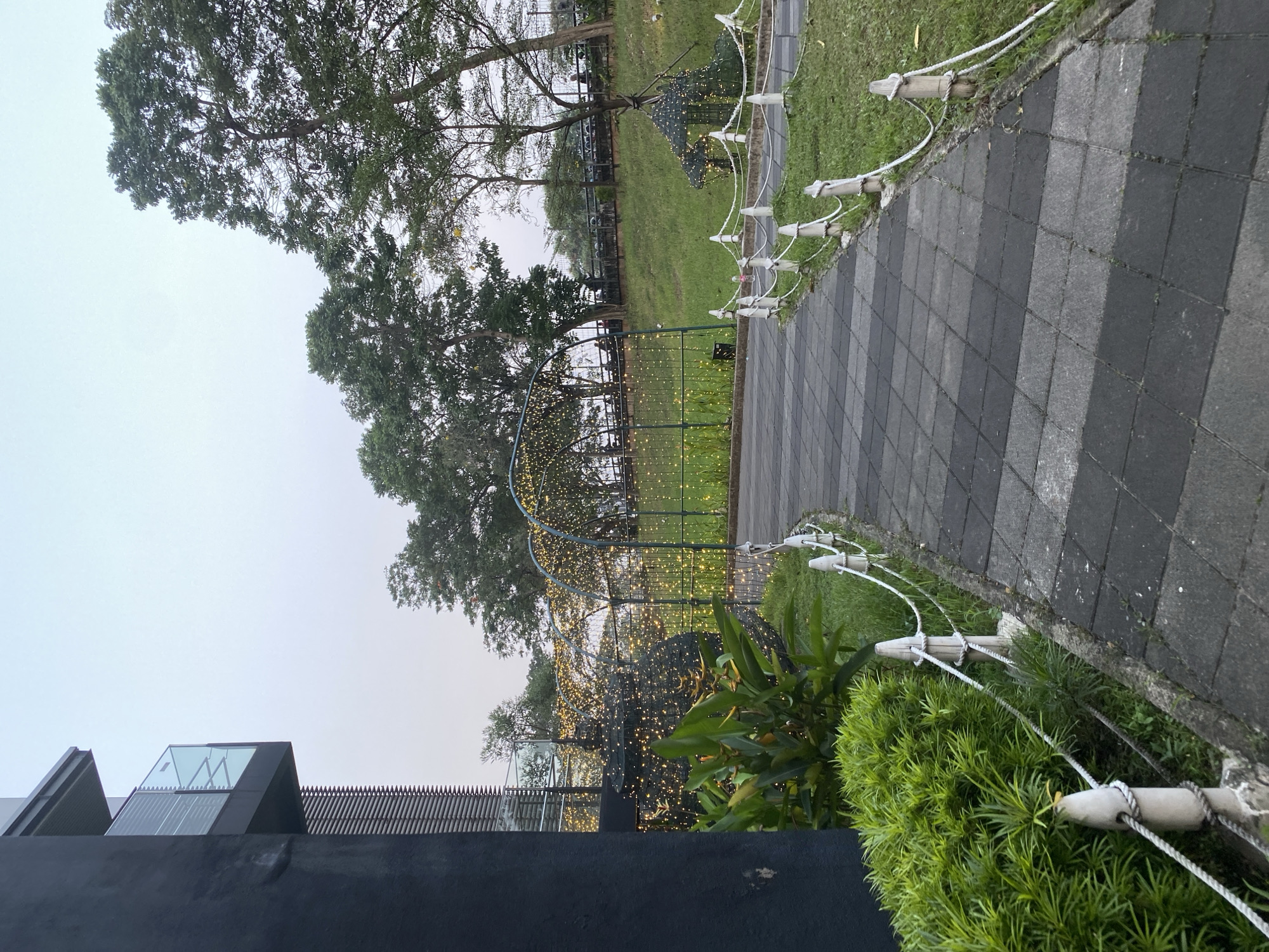 An outdoor pathway in a park area is depicted. The pathway is paved with dark gray and black square tiles, with a white rope and post barrier running alongside. To the left of the pathway, a dark gray building with modern architecture, featuring balconies with glass railings and horizontal louvers, is visible. In front of the building and along the pathway, there is lush green foliage. Beyond the pathway, an archway decorated with numerous small, warm-toned lights is visible. Further into the park, more trees and a grassy slope can be seen, with a small, gazebo-like structure also adorned with lights. The sky is overcast and pale. The scene appears to be in a public park in East Jakarta, Indonesia, possibly at dusk or early morning given the artificial lighting. No people are clearly visible or identifiable in the image.