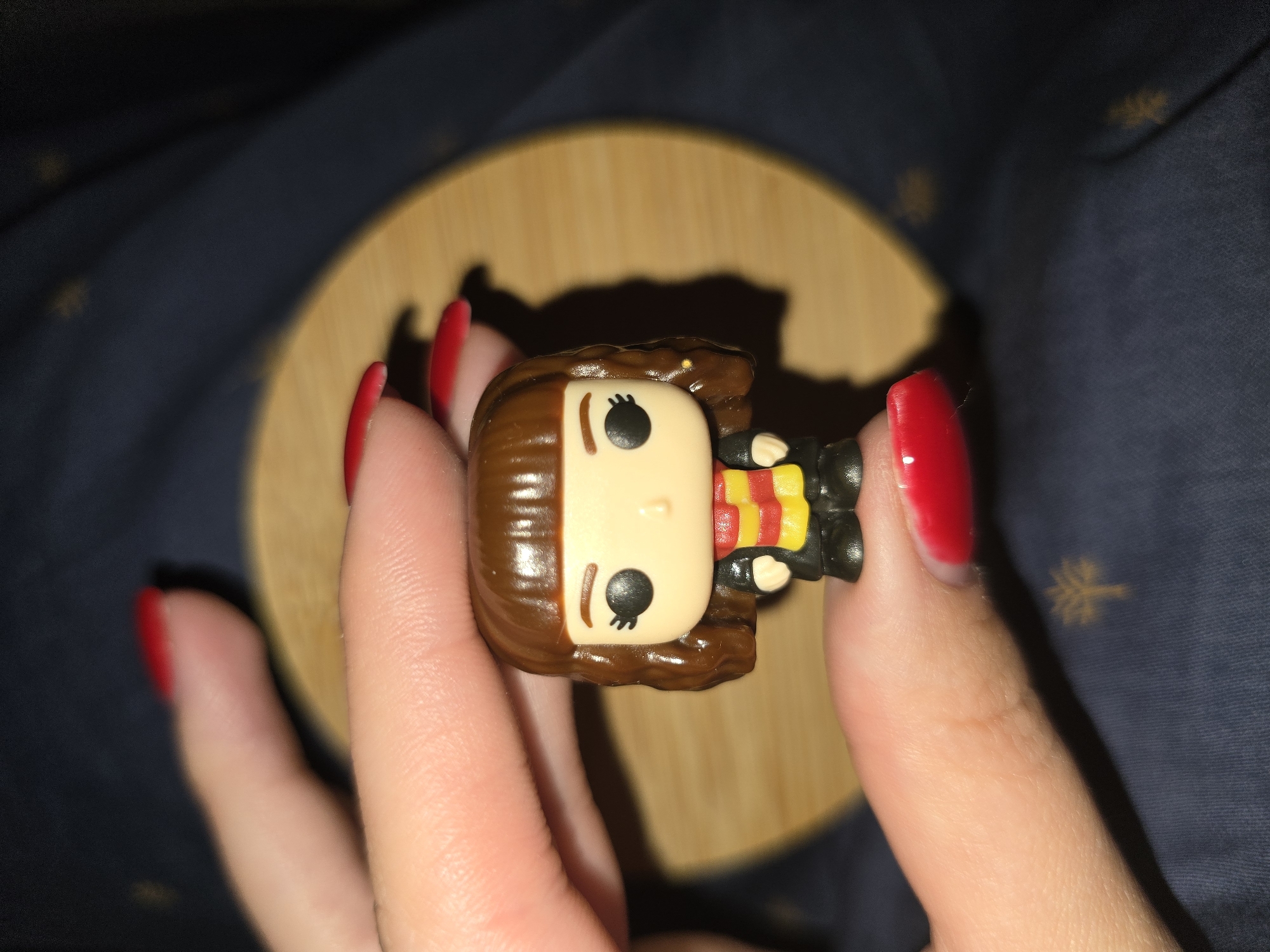 A close-up shot shows a person's hands holding a small figurine. The figurine is a Funko Pop character, likely Hermione Granger from Harry Potter, characterized by her brown hair, pale skin, large black eyes, and a red and yellow striped scarf over a black robe. The figure is held between the thumb and forefinger of the right hand and the index and middle fingers of the left hand. The nails of both hands are painted a vibrant red. The background consists of a dark blue fabric with a subtle pattern and a round, light brown object, possibly a coaster, providing a platform for the figurine. The lighting creates a soft shadow of the figurine and hands on the background. The scene appears to be indoors, with no indication of the time of day or specific weather conditions. No text is visible in the image.