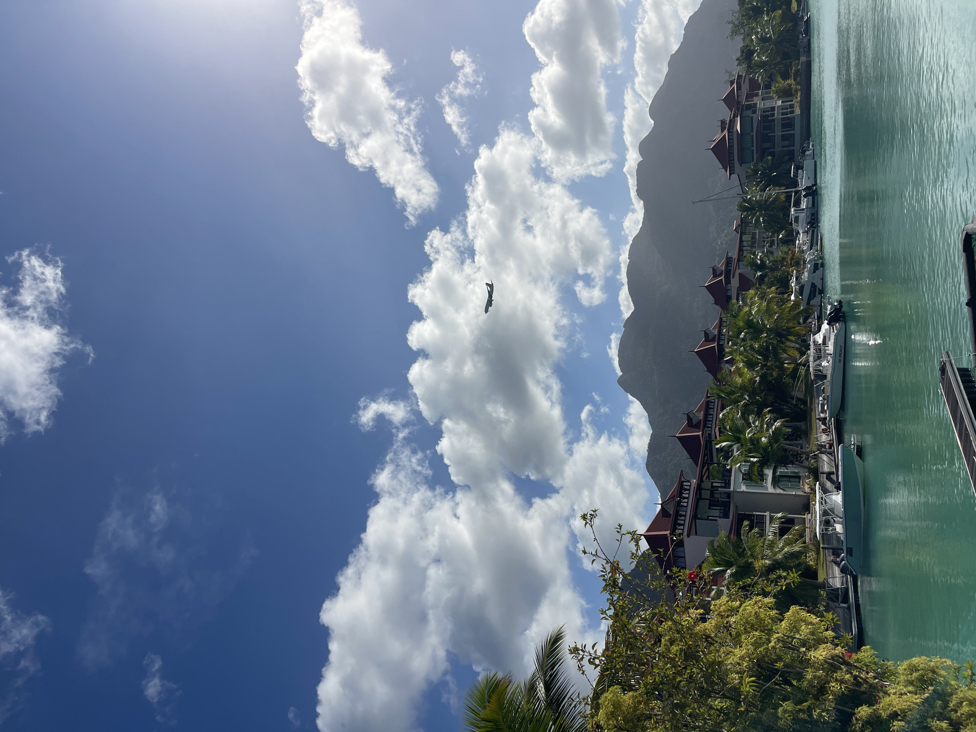 The image depicts a clear blue sky filled with fluffy white clouds, with a single airplane visible in the distance. Below the sky, lush green trees and palm trees frame a scene of coastal architecture. Several multi-story buildings with red tiled roofs and white facades are visible along a waterfront. Numerous boats, ranging from small speedboats to sailboats, are docked in the turquoise water. In the background, a range of majestic, dark, and silhouetted mountains rises against the sky. The overall scene conveys a sense of a tropical resort or residential area in a coastal city. The location is identified as Providence, Seychelles.