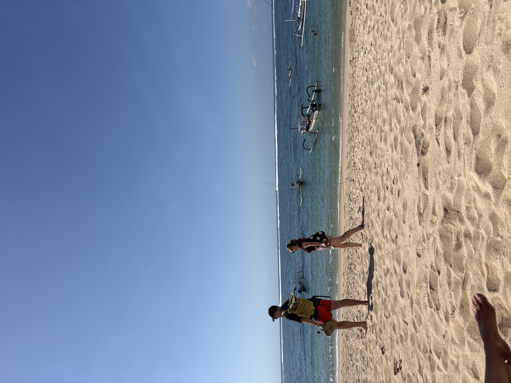 Two people, a man and a woman, walk along the sandy beach. The man, on the left, wears a black t-shirt, red shorts, a black cap, and a bright yellow backpack. He carries a straw hat in his right hand. The woman, on the right, wears a brown crop top, a patterned skirt, and sunglasses, and carries a bag over her left shoulder. The ocean is blue with white waves breaking near the shore. Several traditional Balinese outrigger boats are scattered on the water. The sky is clear and blue. The scene is set in Kuta Selatan, Indonesia, likely on a sunny day.