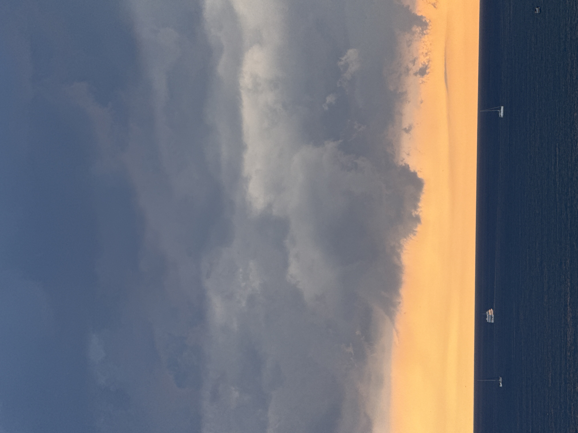 The image depicts a tranquil seascape under a dramatic sky. Dark, heavy clouds dominate the upper portion of the frame, transitioning to a bright orange hue near the horizon, suggesting either sunrise or sunset. Below the sky, the deep blue ocean stretches out, calm and unbroken except for a few distant vessels. On the left side of the image, a white sailboat with a single mast is visible. To its right, a larger white boat, possibly a ferry or a small cruise ship, is also sailing on the water. Further to the right, another small white sailboat is visible. The scene evokes a sense of peace and vastness. This image is unlikely to be from Gelendzhik, Russia, as there are no identifiable landmarks or characteristic features of that city present, and the weather conditions depicted do not align with typical descriptions of Gelendzhik's coastal scenery.