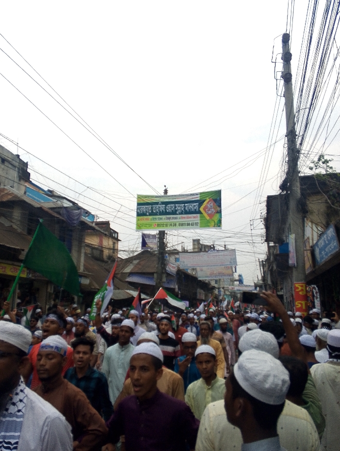 The image shows a large crowd of people gathered in the street in Bhola, Bangladesh, at the coordinates 22.686, 90.644. They are all men, and most are wearing white skullcaps. Some are holding up cellphones and waving flags. Many of the men are wearing long white shirts. There is a large banner hanging above the crowd that is written in Bengali. In the background there are buildings lining the street. The sky is cloudy and there are many power lines strung across the sky. The crowd appears to be a religious procession.