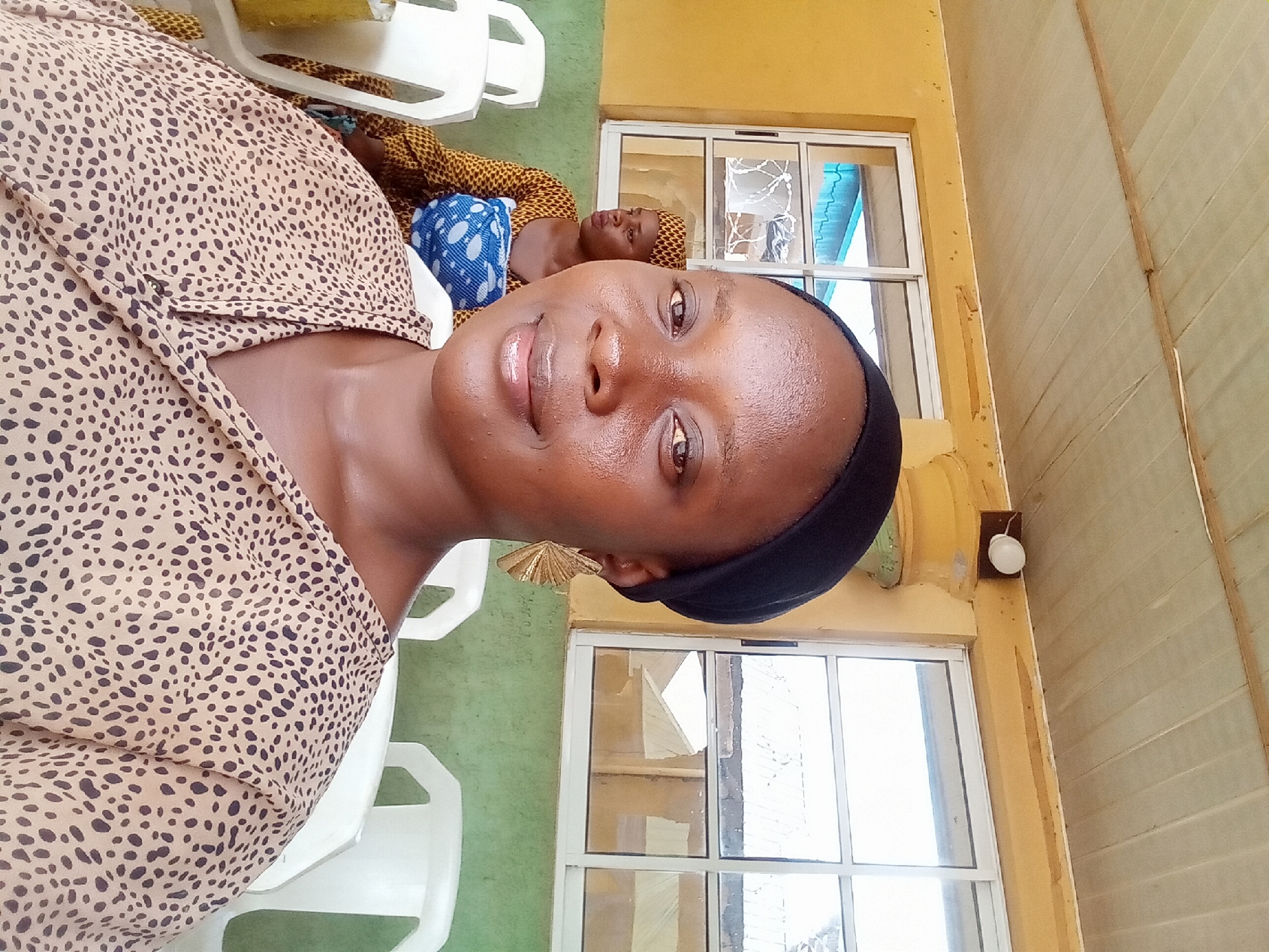 A medium close-up shot, taken in Rido, Nigeria, presents a smiling woman in a brown, patterned shirt and a black head covering. She appears indoors, seated on a white plastic chair. Behind her, another person rests on what appears to be another chair, covered with a colorful fabric and a blue and white headscarf. The setting includes a yellow-painted wall with two white-framed windows. A light fixture is noticeable on the wall to the right of the windows. The environment gives the impression of a modest indoor space.