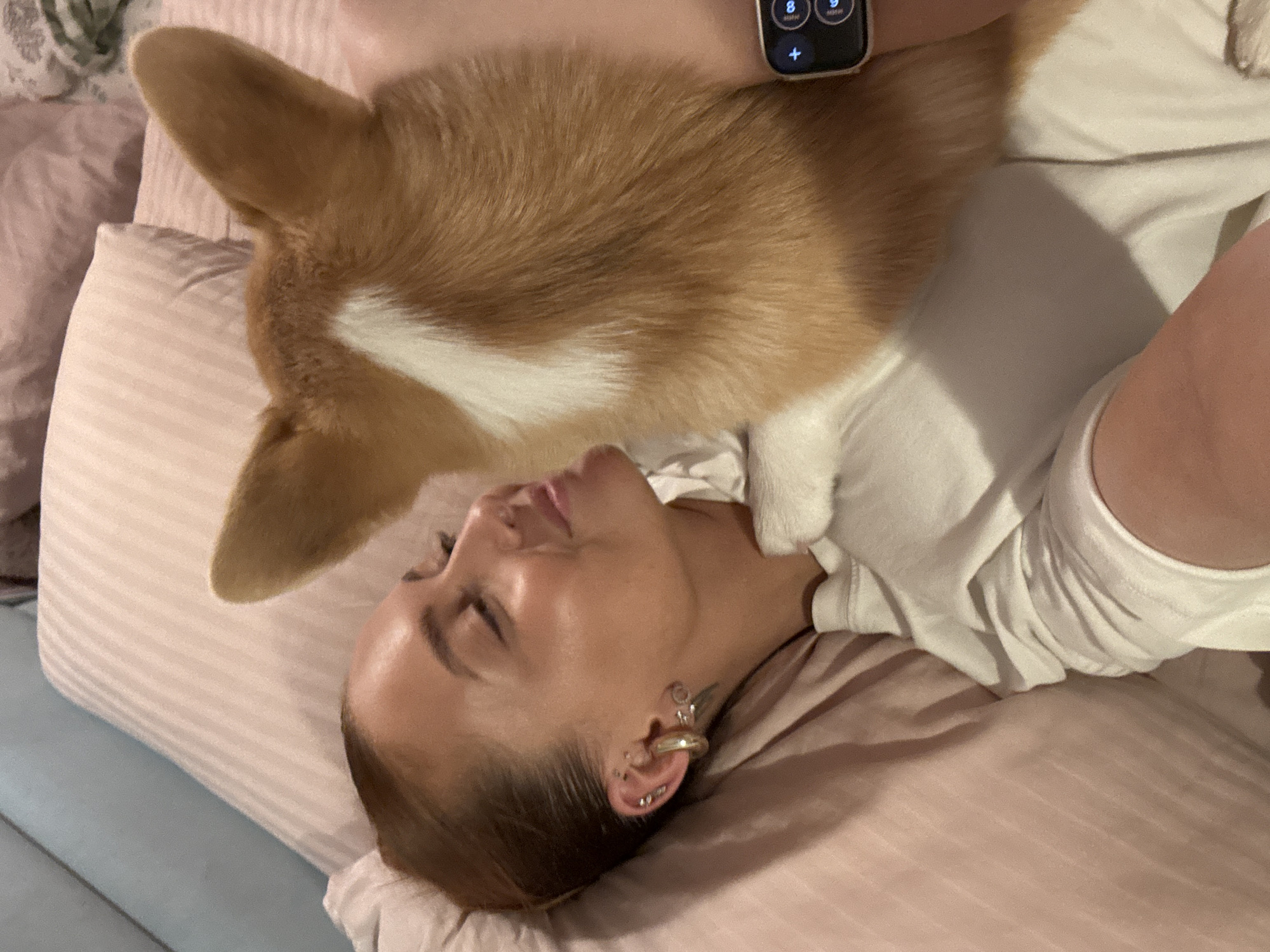 A woman is lying on a pink bed with a corgi dog lying on top of her. The woman is wearing a white top and has several piercings in her left ear. She also has a smart watch on her left wrist. The dog is laying on the woman's neck. The background is blurred. There is no visible evidence to suggest this image was taken in Chaikovsky, Russia.