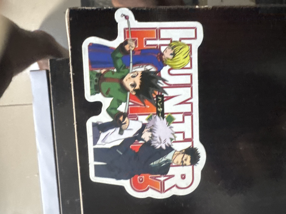 The image presents a rectangular sticker affixed to a dark, flat surface, possibly a box or shelf, located in Ipetumodu, Nigeria. The sticker depicts four anime characters from "Hunter x Hunter" standing in a line, with the series title "HUNTER HxH" displayed in large red and white lettering, accompanied by smaller Japanese text. From left to right, the characters are: a blond-haired individual in a blue tunic; a dark-haired character in a green outfit, holding a fishing rod horizontally across his shoulders; a white-haired character in a white shirt; and a dark-haired character wearing glasses and a dark suit. The dark surface shows minor surface wear. Below the surface, light-colored floor tiles are visible, and a blurred, dark-skinned human foot is partially present in the lower left foreground.