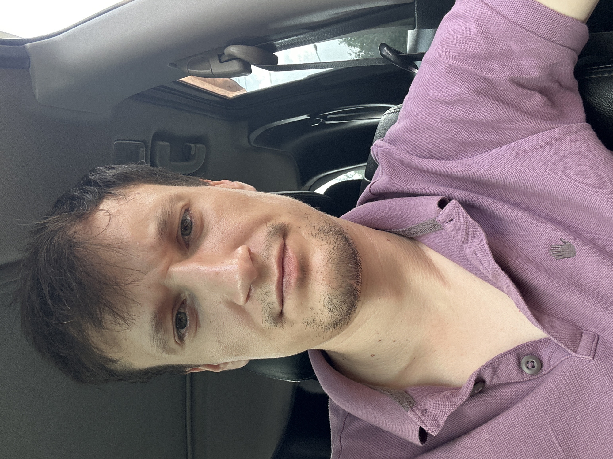 A man is seated in the driver's seat of a car, taking a selfie. He has light brown hair and stubble. He is wearing a purple polo shirt with a gray collar. The interior of the car is visible, including the dashboard and seatbelt. The background is blurred, but appears to be outdoors, possibly in a residential area. The scene is lit by natural daylight. The image does not provide any specific location information within the city of Makiv, Ukraine, or any other specific geographical markers.