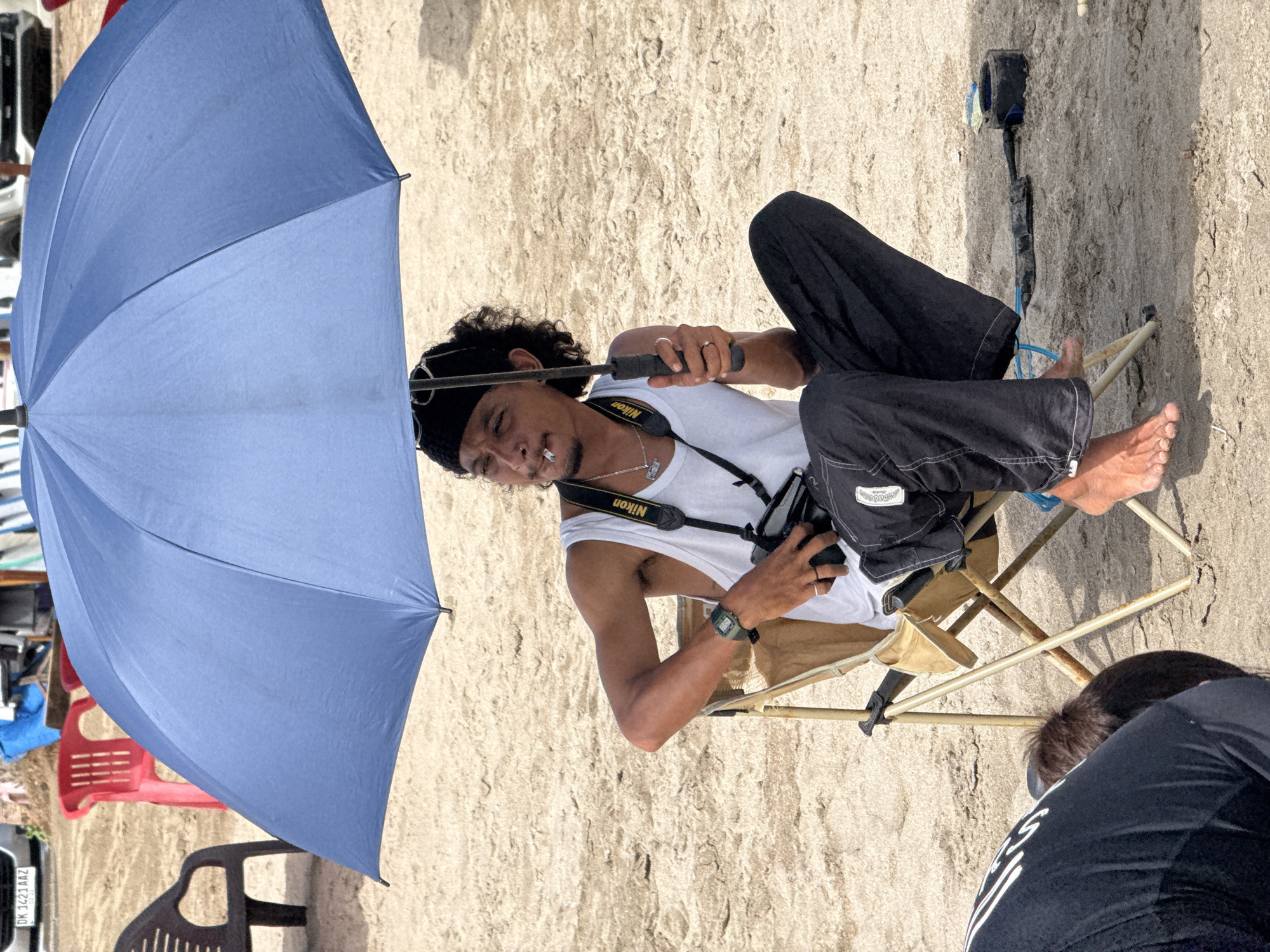 The image shows a man sitting in a beige folding chair on a sandy beach. He is holding a blue umbrella. He has curly black hair, a black bandana, a white tank top, and dark pants. A Nikon camera strap is around his neck. A cigarette is in his mouth. The beach sand is light brown and there are other people and objects such as a plastic chair, a car and a blue tarp in the background. There's a surf leash next to him on the sand. Another person is partially visible in the lower right corner of the image. The scene is likely in Kuta, Indonesia, given the presence of a license plate with the letters "DK" and a series of numbers and letters.