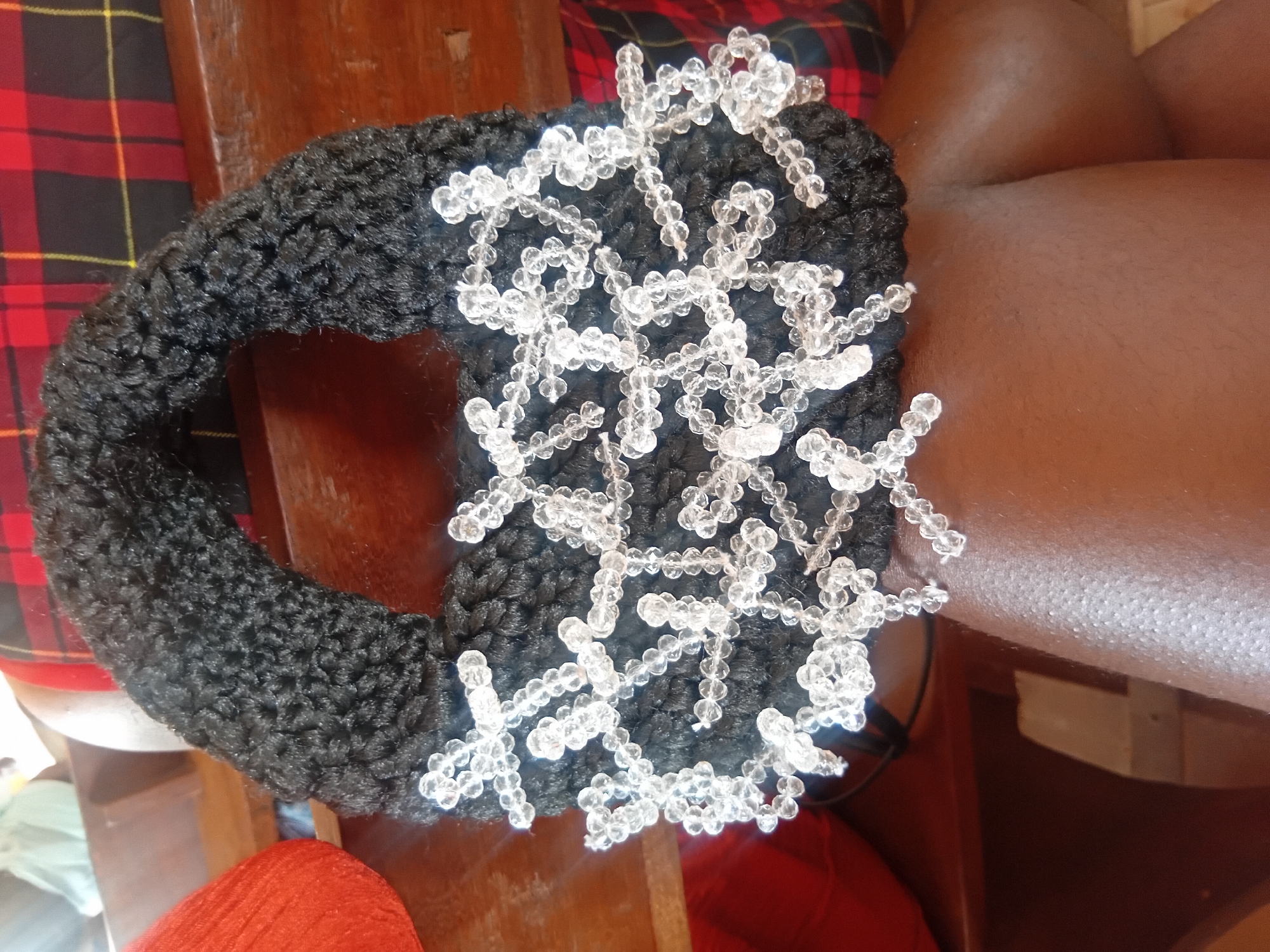 The image shows a close-up of a handmade black crocheted bag with clear beaded embellishments, resting on a person's leg. The bag has a rounded shape and a handle. The beads are arranged in a delicate, intricate pattern across the front of the bag. In the background, a wooden surface is visible, along with a red and black plaid fabric. The setting appears to be an indoor or semi-outdoor environment, potentially a home or a market stall. The overall impression is one of craftsmanship and a personal item being displayed or held. There are no discernible landmarks indicating the specific location in Ejule, Nigeria.