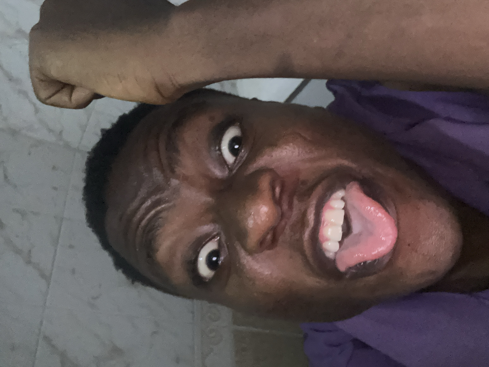 A close-up, slightly angled, indoor photograph shows a dark-skinned person with their tongue sticking out. Their eyes are wide. They are wearing a purple garment. The person has short, dark hair. A clenched fist is visible above their head. The background appears to be a tiled wall. No specific details point to Ikorodu, Nigeria.