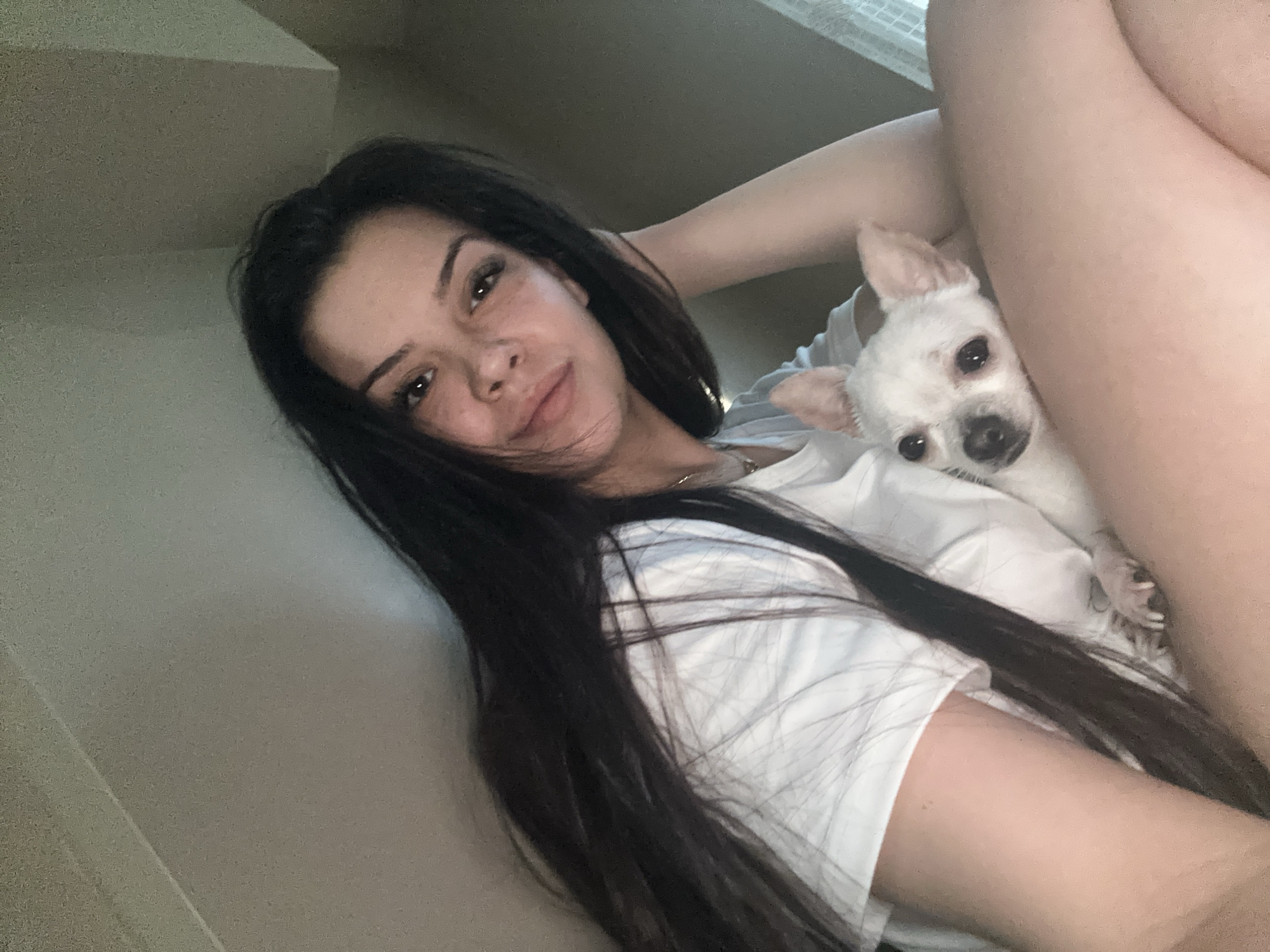 This portrait-oriented image features a young woman and a small white dog in an indoor setting, presumably in Sliven, Bulgaria. The woman, with long, dark hair flowing over her shoulders, is seen from a low angle, looking directly at the camera with a relaxed, gentle smile. Her face shows natural skin texture, possibly freckles, and she wears a simple white t-shirt and a thin necklace. Her right arm is bent, with her hand near her head, and her bare legs are visible in the upper right of the frame, suggesting she is lounging or reclining.
Nestled closely against her chest is a small, white dog, resembling a Chihuahua, with large ears, dark eyes, and a dark nose. The dog is also looking towards the camera, appearing calm and comfortable. The background is indistinct, showing light-colored walls or a ceiling, typical of an interior space. Soft, natural light illuminates the scene, suggesting it is daytime. The overall impression is one of affectionate companionship and relaxation. No text is visible in the image.