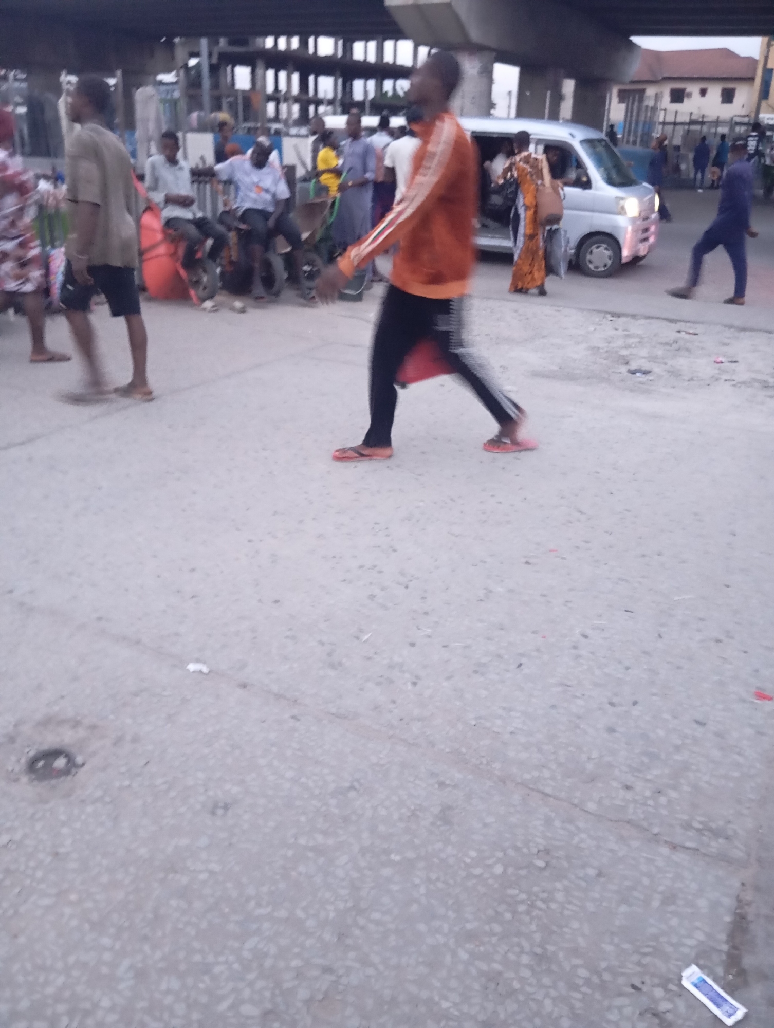 A busy street scene unfolds under an elevated highway structure in Port Harcourt, Nigeria. In the foreground, a man in an orange and black tracksuit walks past, his form blurred by motion. His feet are adorned with bright orange flip-flops. To his left, a man in a light brown shirt and dark shorts walks away from the camera. Further back, a cluster of people and vehicles indicates commercial activity. A silver minivan is parked, with individuals entering and exiting. Several motorcycles and a red cargo bag are visible near a group of seated individuals. The environment appears to be a paved street with some debris scattered on the ground. The overall impression is one of a dynamic urban environment with constant movement and pedestrian and vehicular traffic.