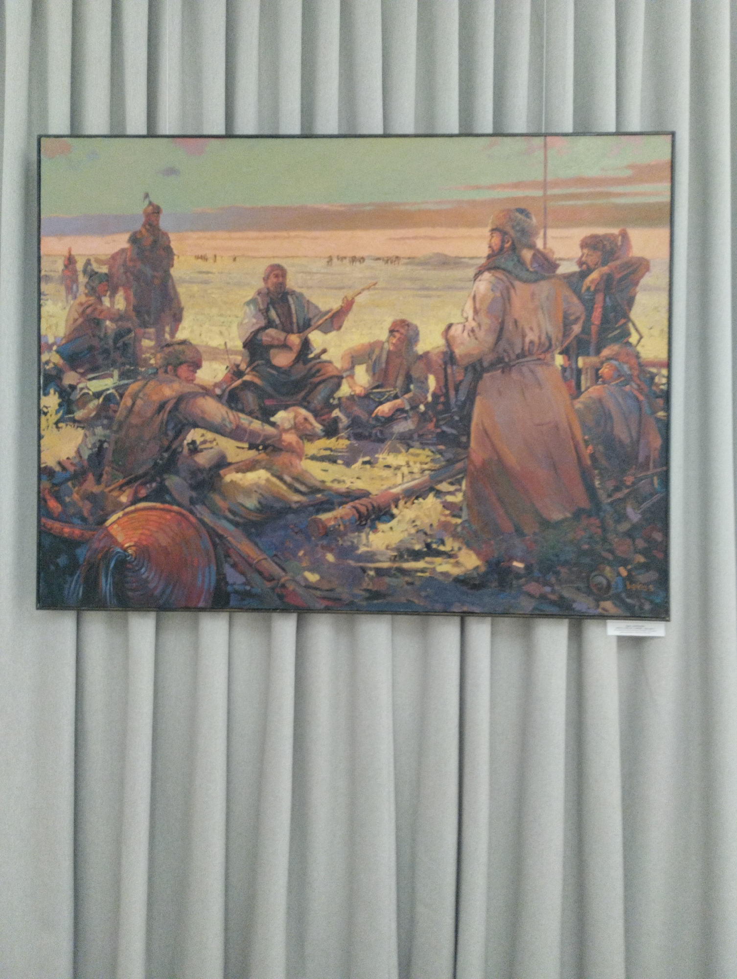 The image shows a painting depicting a group of people in what appears to be a historical or cultural setting, likely in Kazakhstan, given the location context. The painting features several individuals, some on horseback and others gathered around a fire or a campsite. The figures are dressed in traditional clothing, including fur hats and long robes, suggesting a nomadic or historical theme. One person in the center is playing a stringed instrument, perhaps a dombra, indicating a musical or social gathering. Other individuals are engaged in various activities, such as tending to a fire or interacting with each other.
The scene is set in an open, arid landscape with a muted color palette, suggesting either dawn or dusk. The sky is painted with soft hues of orange and yellow, hinting at a warm atmosphere. The overall mood of the painting seems to be one of communal gathering and storytelling, with a sense of historical significance. There are also some objects visible, including what might be a saddle and other equipment, further reinforcing the historical and nomadic theme. There is a small white label attached to the bottom right of the frame, but the text is not legible. The painting is framed and hung against a backdrop of grey vertical blinds.
