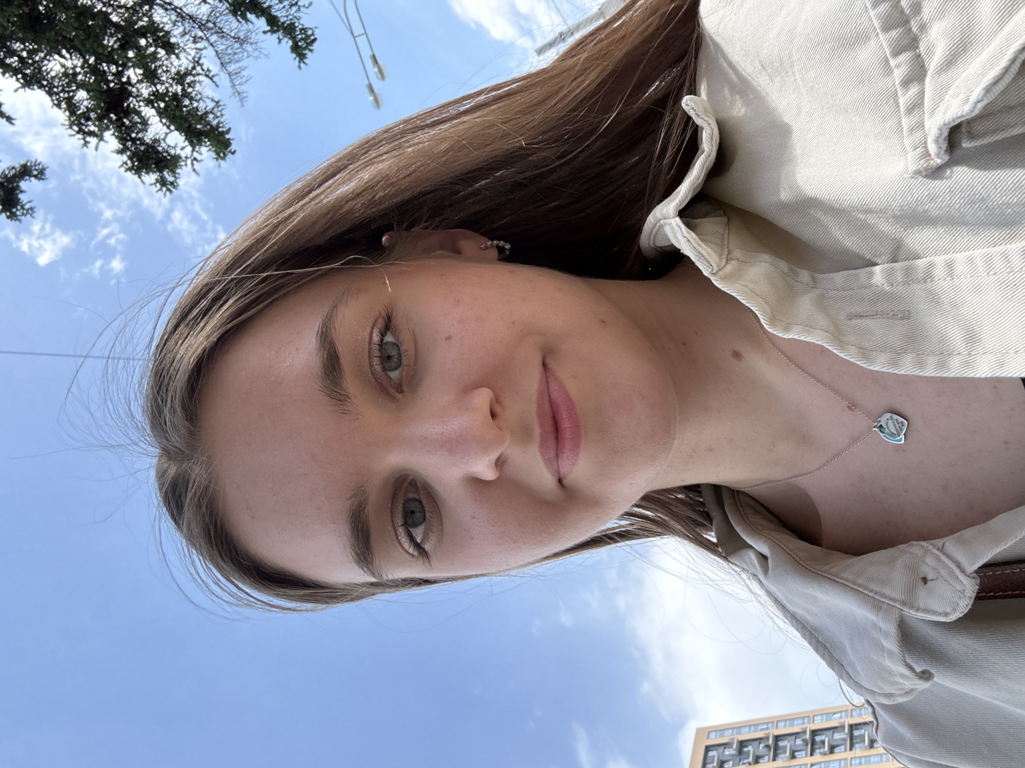 A selfie taken from a low angle. The subject, a young woman with light brown hair, light skin, and light green eyes, is centrally framed. She is wearing a light-colored jacket and a necklace with a heart-shaped pendant. Behind her is a bright blue sky with some white clouds, and in the background, trees and a tall, light-colored building with many windows can be seen. The photograph appears to have been taken in an urban environment, in the city of Perm, Russia.