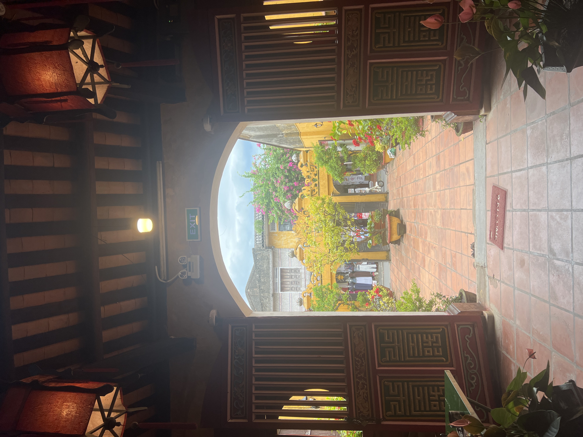 The image shows an interior view looking out through an arched doorway into a courtyard in Hoi An, Vietnam. The doorway is flanked by two large, intricately carved wooden doors, which are partially open. Above the doorway, a sign with the word "EXIT" is visible. Hanging lanterns, one on each side, are illuminated and add to the ambiance.
The courtyard is paved with terracotta tiles and is filled with potted plants and flowers, adding vibrant splashes of color. In the background, yellow buildings with decorative architectural details are visible, characteristic of Hoi An's historic architecture. Various items, possibly for sale or display, are arranged in the courtyard, suggesting a market or shop area. People are present in the courtyard, some appearing to be browsing or tending to the stalls. The sky above is partly cloudy. A red "WELCOME" mat is placed on the tiled floor in the foreground.