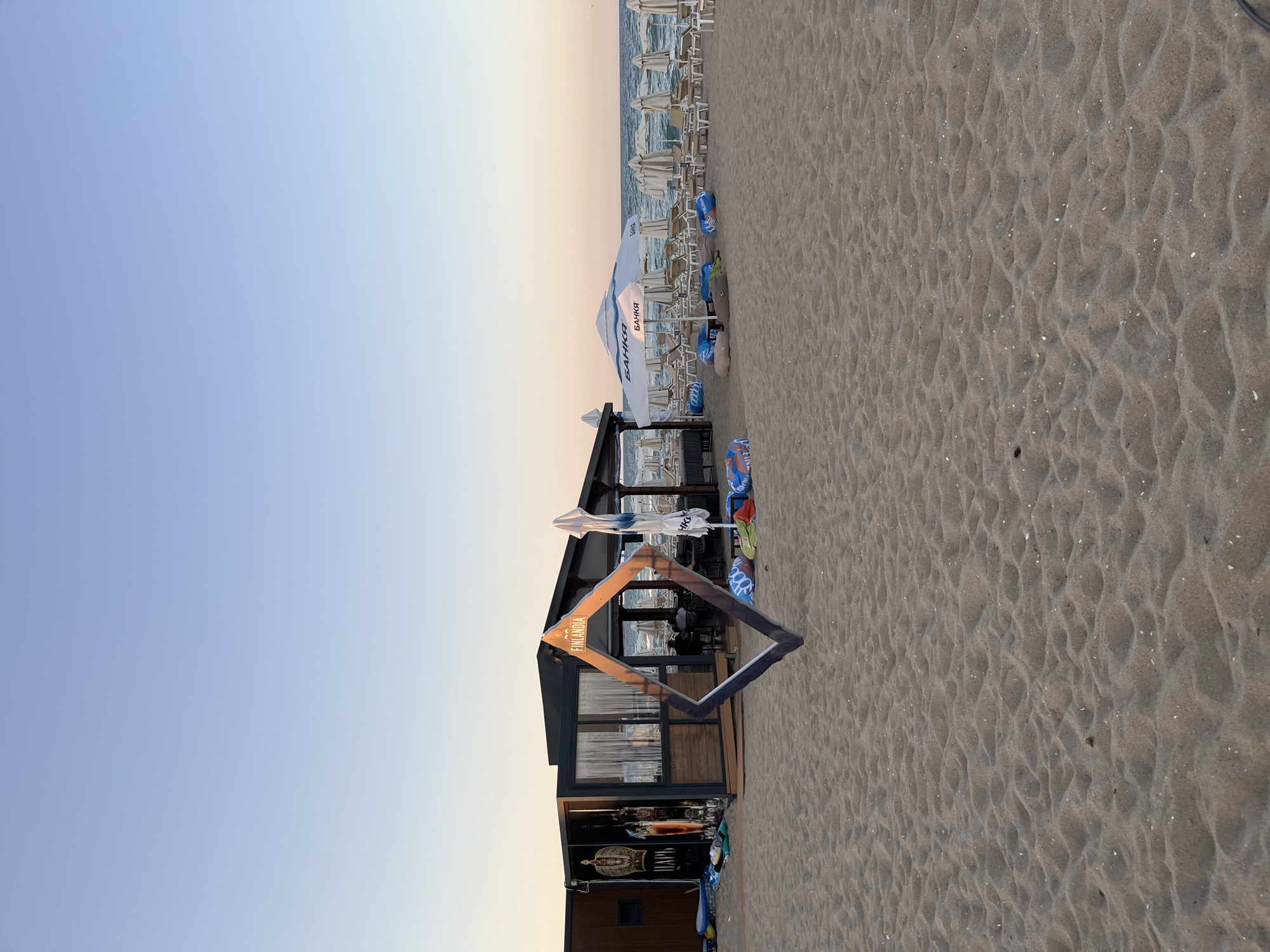 A beach scene in Obzor, Bulgaria, shows a lounge area with beach chairs and umbrellas set up on the sand. A wooden structure, possibly a bar or cafe, is visible on the left, with a large diamond-shaped frame positioned in the foreground. The sky is clear and blue, with the ocean stretching out in the background. The beach is mostly empty, with no people in sight.