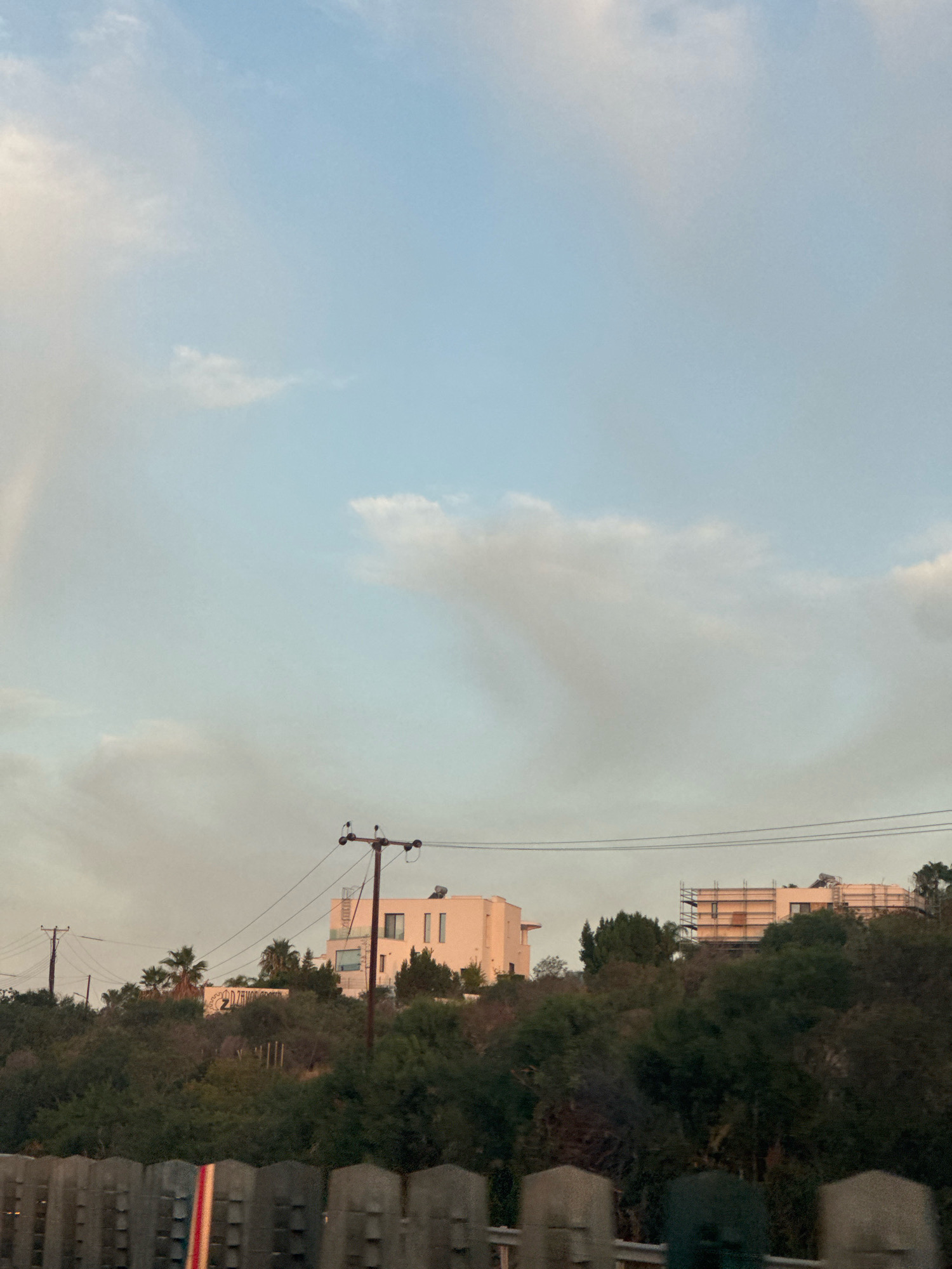 A residential area on a hillside in Mouttagiaka Community, Cyprus, is depicted in this image. The scene is dominated by a light blue sky with scattered clouds. In the middle ground, a cluster of buildings and trees are visible on the elevated terrain. Two distinct buildings are prominent: one is a light-colored, multi-story structure with several windows, and the other appears to be under construction, indicated by scaffolding. Telephone poles with wires run across the scene, suggesting utility infrastructure. The foreground is partially obscured by a wooden fence with vertical slats. The overall environment suggests an outdoor, daytime setting, possibly taken from a road.