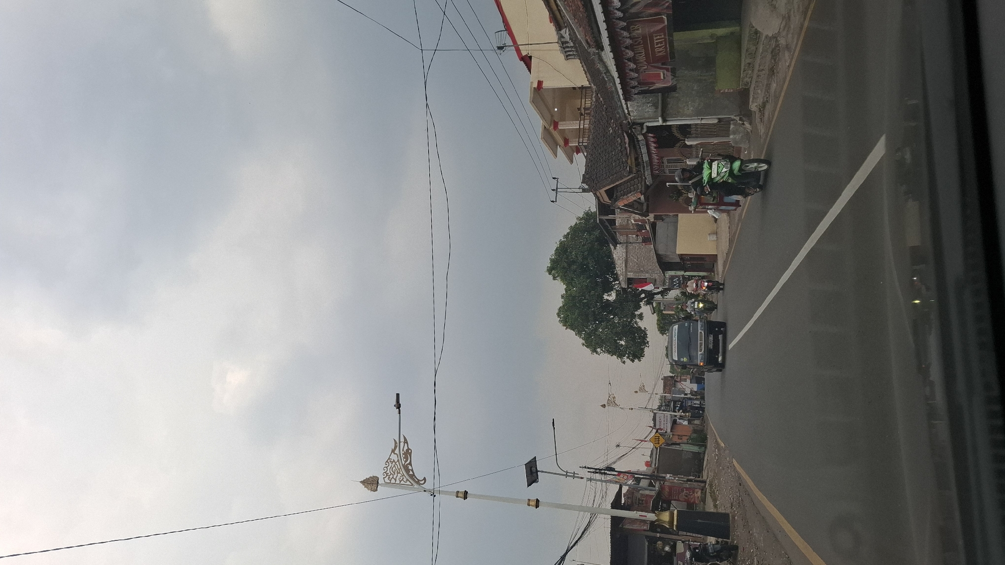The image depicts a street scene in Cilaku, Indonesia, under an overcast sky. A paved road with a white and yellow line marking runs through the center of the frame. Several vehicles are visible on the road, including a dark-colored van, three motorcycles, and a green scooter with a passenger. On the left side of the road, there are buildings with shops and what appear to be streetlights with decorative elements. On the right side, more buildings are present, including houses and a shop with signage. A large tree with dense foliage stands between the buildings. The overall impression is a typical residential and commercial street in a town or city.