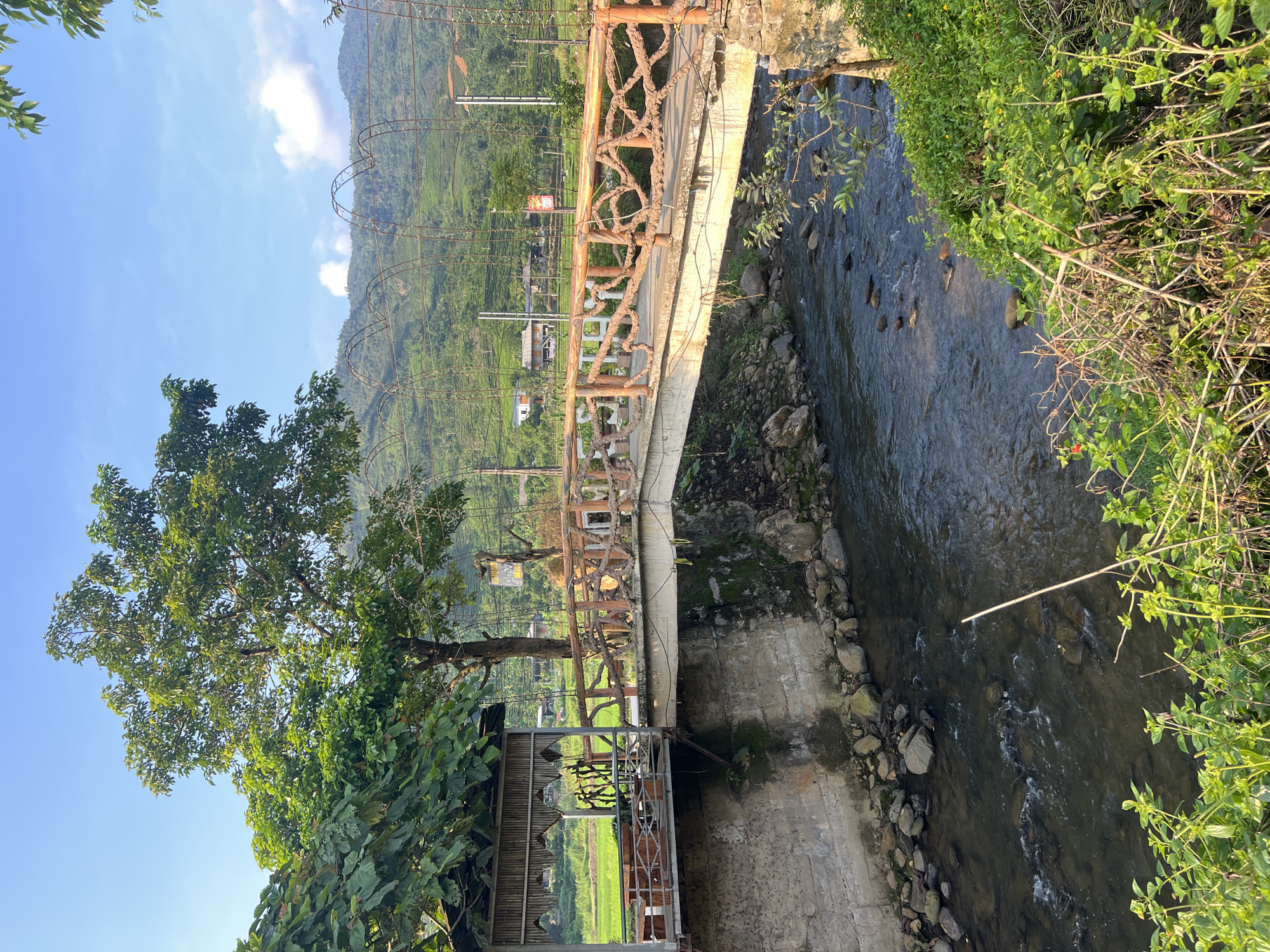 The image shows a bridge over a shallow river in Gia Hội, Vietnam. The bridge is made of concrete with a decorative wooden railing featuring intertwined branches. To the left of the bridge is a shaded seating area with bamboo walls and a thatched roof, overlooking a vibrant green field. A large tree with lush foliage stands behind the seating area, casting shadows. The riverbed is visible, with rocks scattered throughout the clear, flowing water. In the background, rolling hills covered in terraced vegetation stretch towards a bright blue sky with scattered clouds. The scene depicts a tranquil natural environment with some man-made structures.