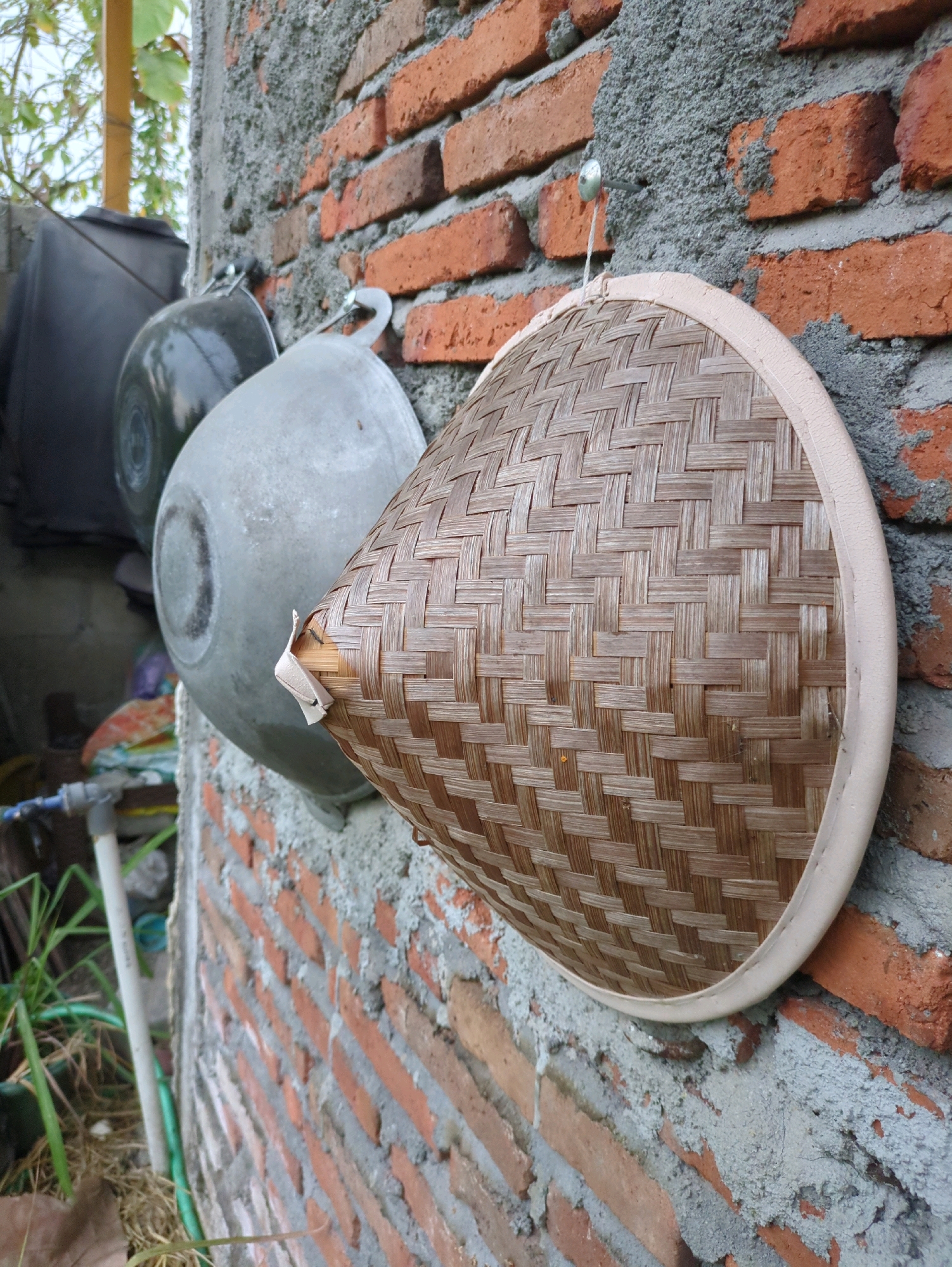 In Sindon, Indonesia, a woven bamboo conical lid, approximately 30 cm in diameter, hangs on a nail against a rough brick wall. The lid is bordered by a cream-colored fabric. Behind it, two metal pots, one dark gray and the other light gray, are also hung on the wall. To the left, a dark plastic container is partially visible. The wall itself is constructed of red bricks with visible gray mortar. In the lower left corner, a white pipe with a blue fitting and a green hose are visible, surrounded by dry grass and leaves. The scene appears to be outdoors, possibly in a backyard or storage area.
